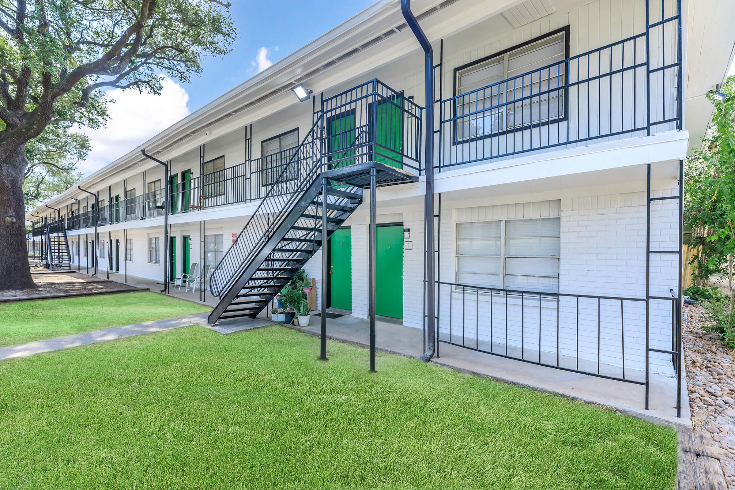 A view of a two-story residential building with a green and white color scheme. It features a metal spiral staircase leading to the upper floor, green doors, and a well-maintained grassy area in front, with trees lining the side of the building.
