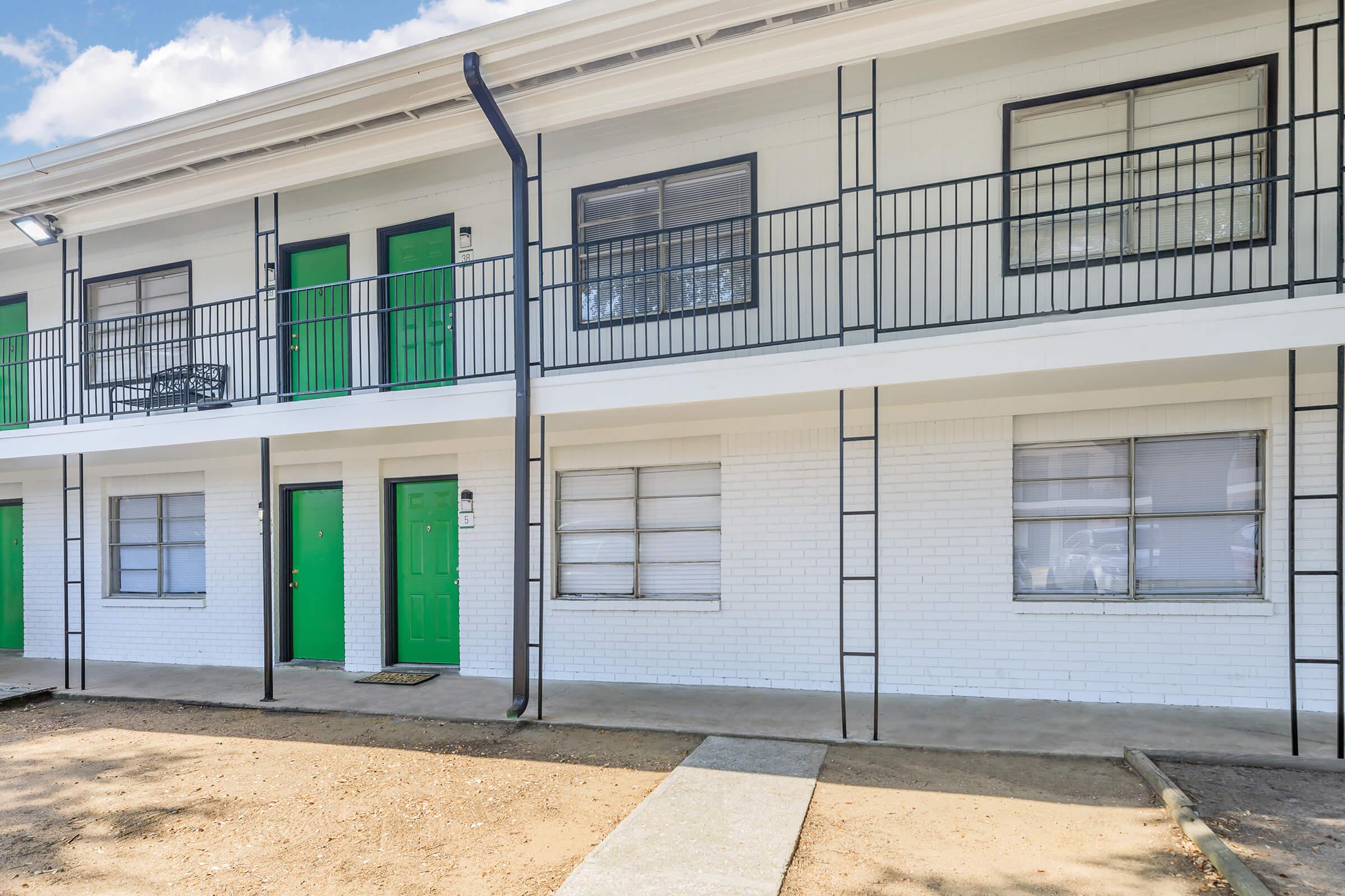 Exterior view of a two-story building featuring white brick walls, black railings, and bright green doors. The ground is a gravel or dirt surface, and there are windows with white blinds. The setting appears to be a residential or motel-style structure with a simple and clean design.