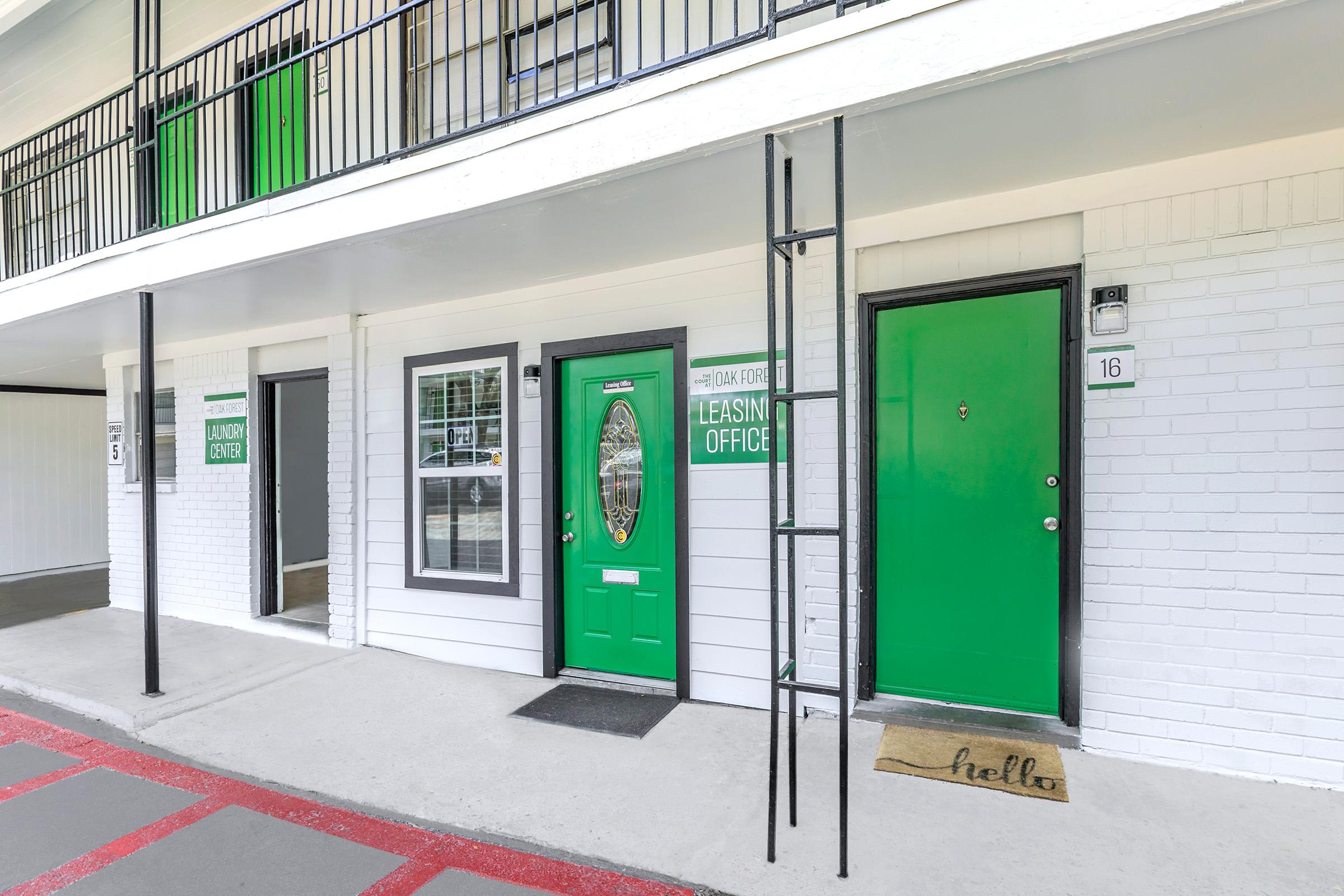 A view of an apartment building's exterior with three doors. The central door, painted green, is labeled "Leasing Office." To the left is a door marked "Laundry Room," and to the right is a green door labeled "16." A black metal ladder leans against the wall, and a welcome mat reads "hello."