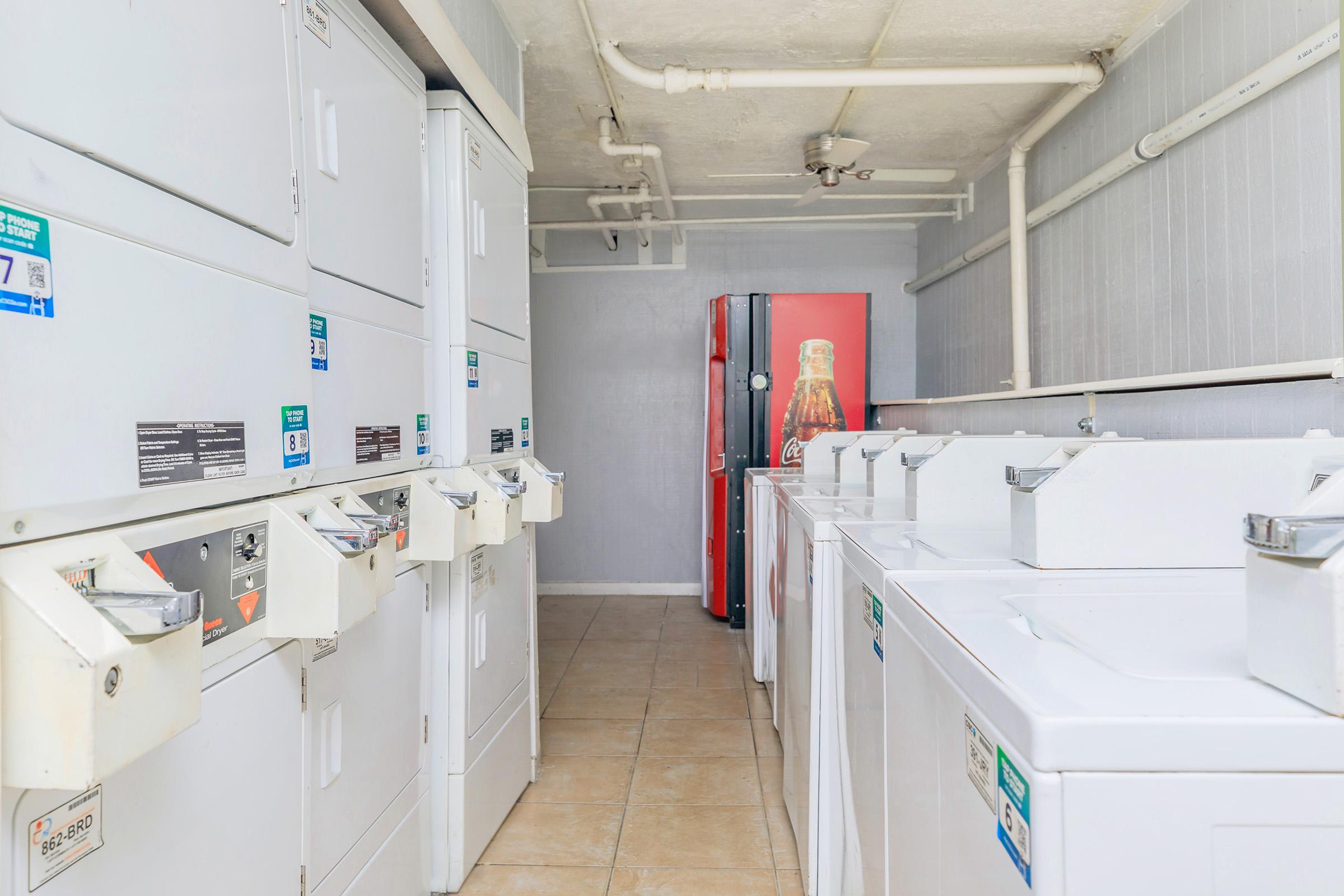 A clean laundry room featuring several white washing machines and dryers lined up along the walls. There are coin-operated payment stations on each machine. In the background, a red refrigerator and a poster of a soda can be seen, adding color to the space. The room has tiled flooring and neutral-painted walls.