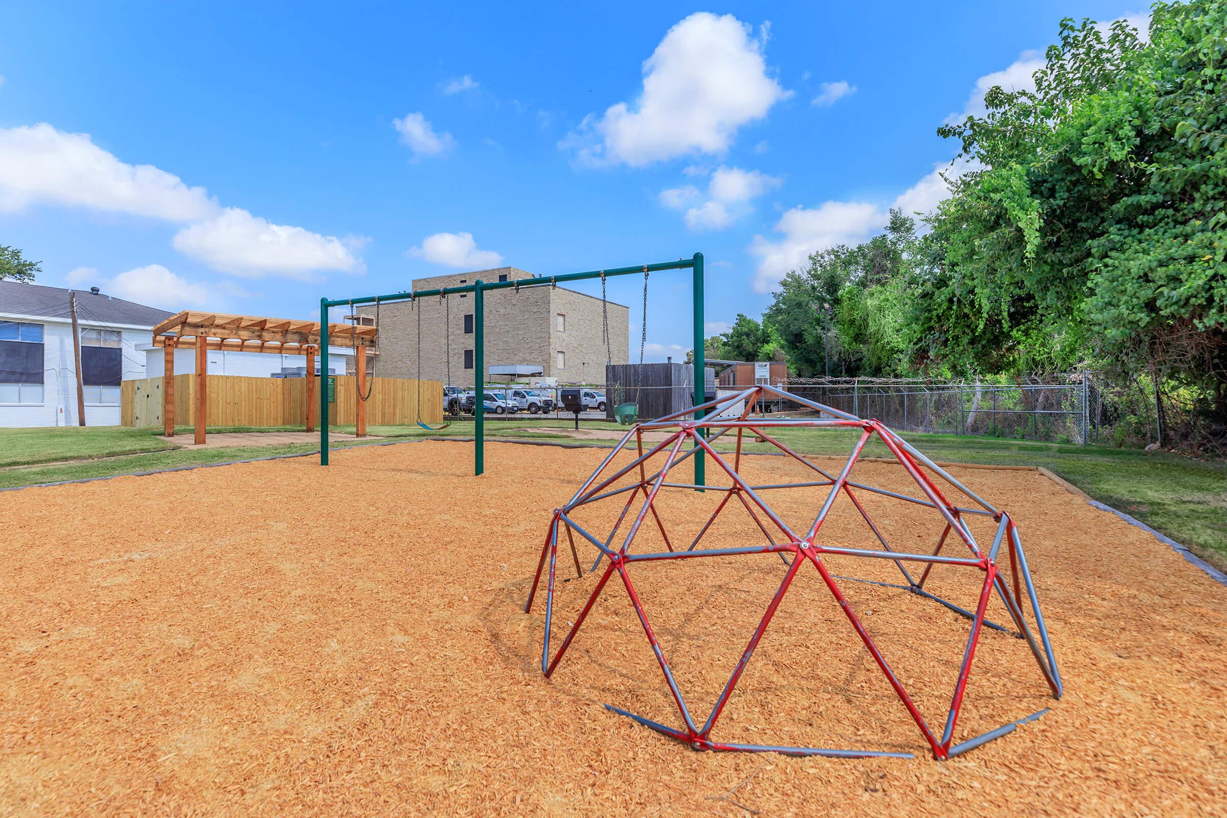 A playground featuring a geodesic climbing dome and swings, surrounded by a grassy area and a wooden fence. In the background, there is a building and several parked cars under a clear blue sky with scattered clouds.