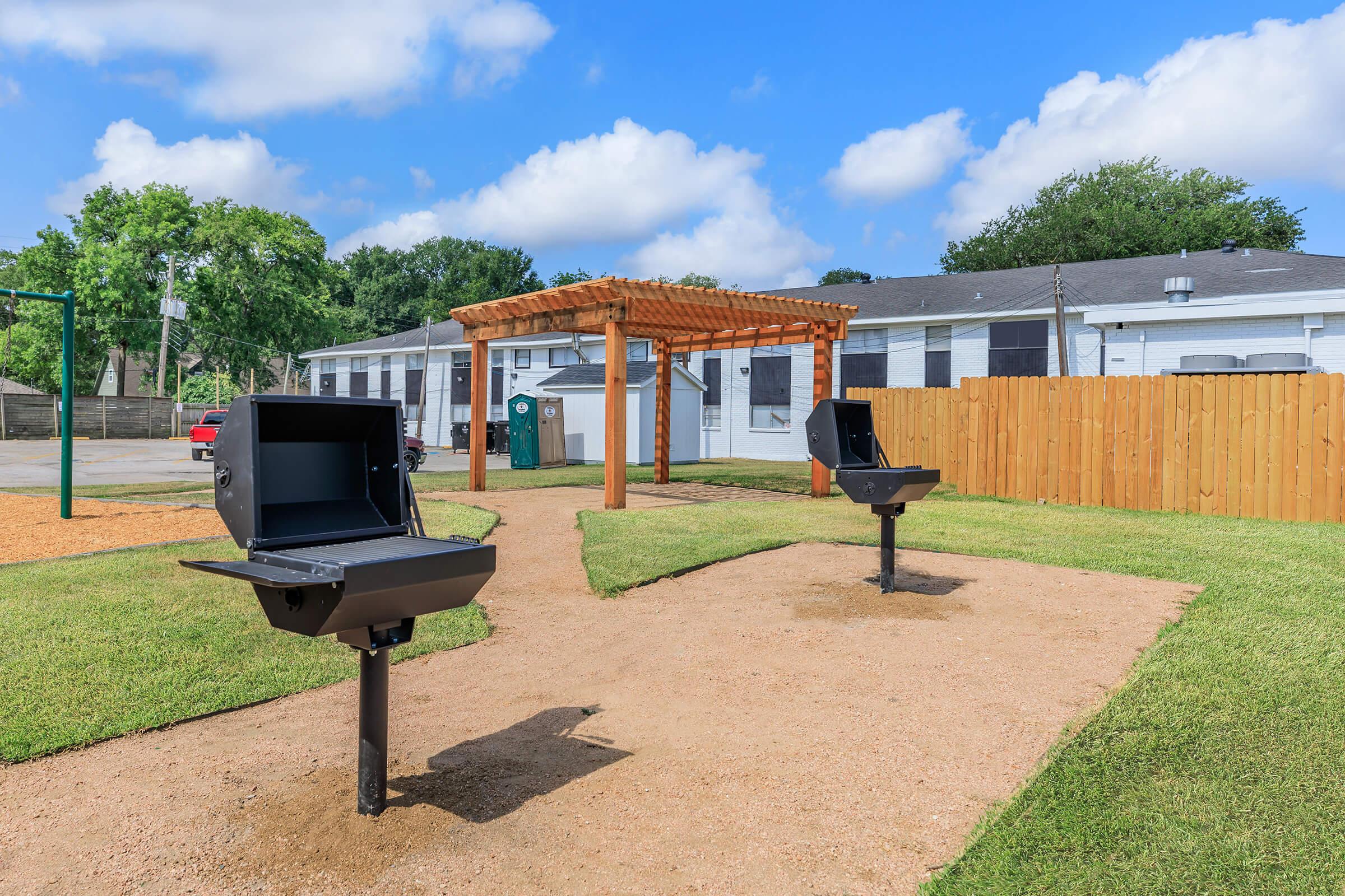 Two charcoal grills are set up on a gravel pathway in a park-like setting. A wooden pergola is in the background, surrounded by grassy areas and a fence. There are trees and a blue sky with clouds, creating a welcoming outdoor space for gatherings and barbecues.