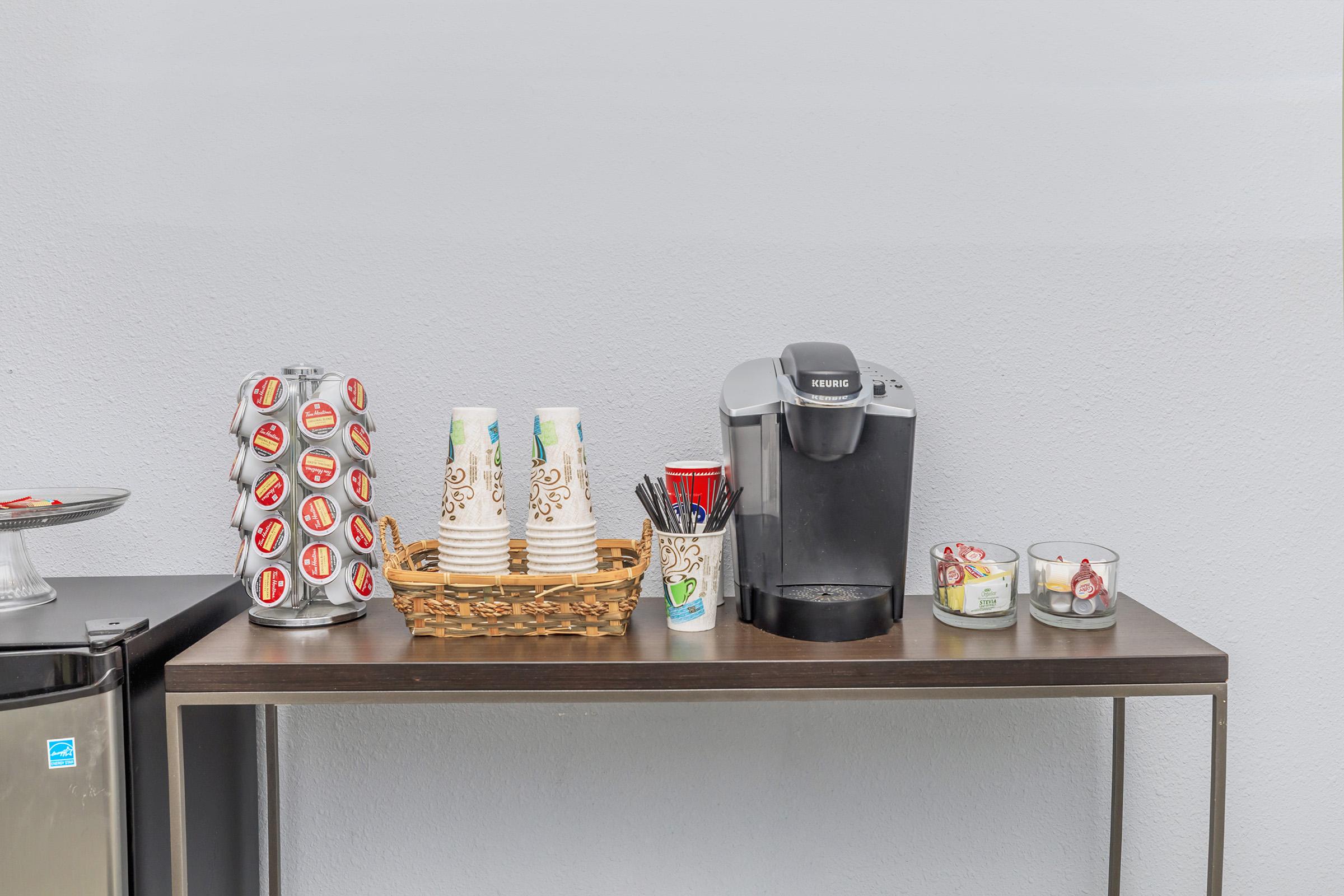A coffee station featuring a Keurig machine, a basket of disposable coffee cups, a tower of coffee pods, and containers of stirrers and sugar. There are also small candles on the table, set against a simple gray wall.