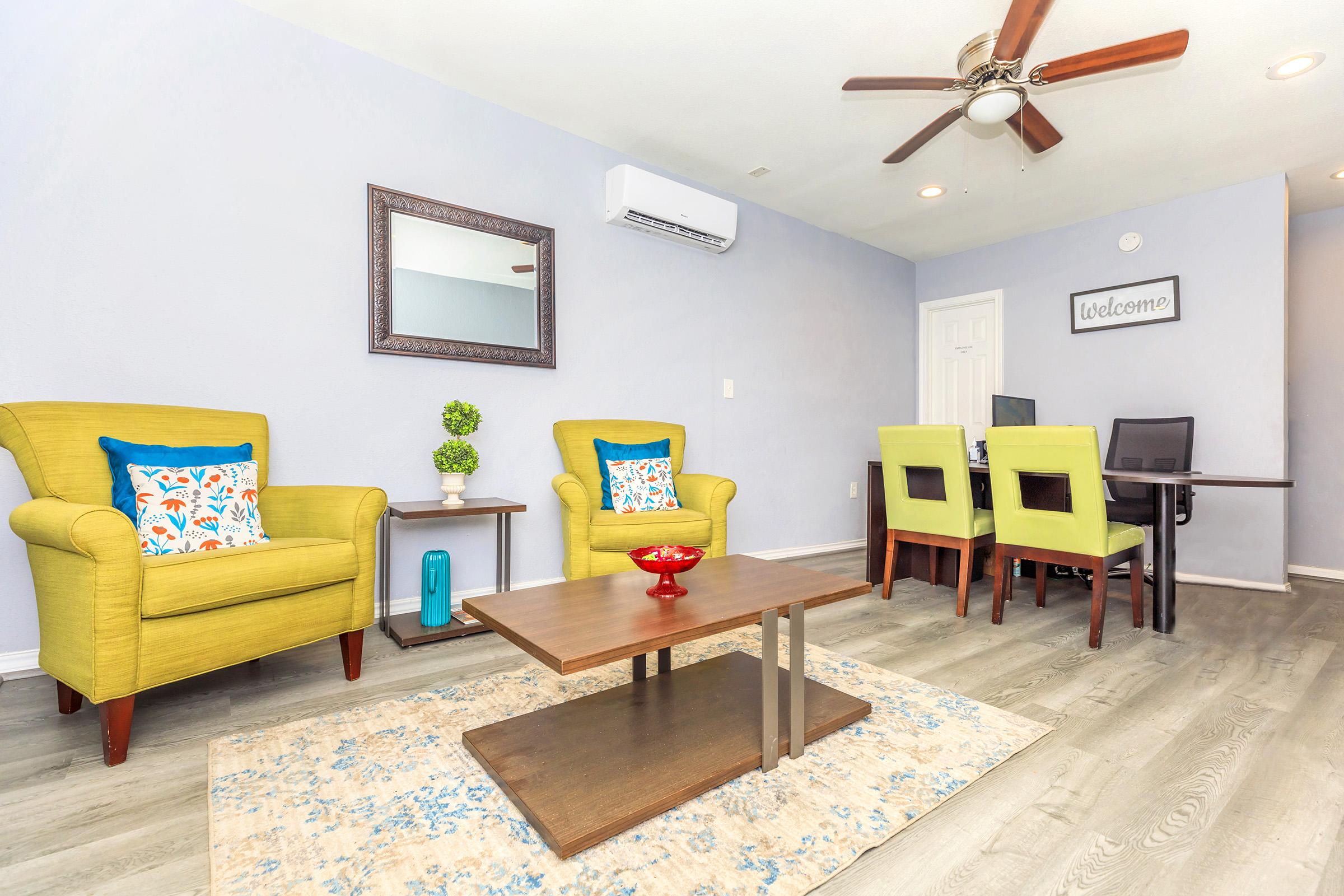 Interior of a modern living room featuring two bright yellow armchairs with colorful cushions, a wooden coffee table, and a small decorative plant. In the background, there's a welcome sign, a mirror, and a dining area with a dark table and chairs. The walls are painted light blue, and the flooring is light gray wood.