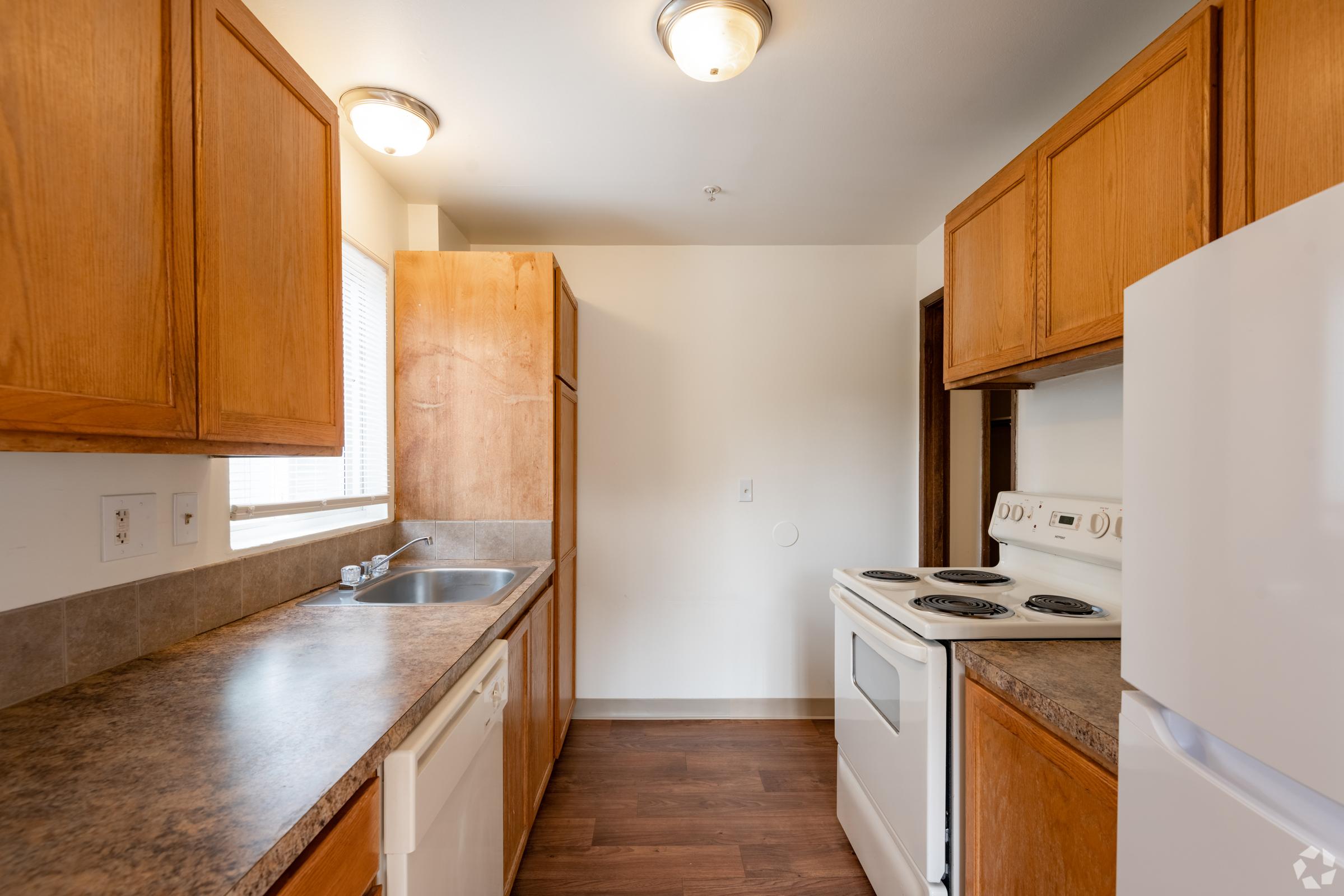 A modern kitchen featuring wooden cabinets, a white stove, and a dishwasher. The countertop is dark, and the flooring is a warm brown wood. Natural light enters through a window, illuminating the space. The layout is open, with ample storage and a functional design.