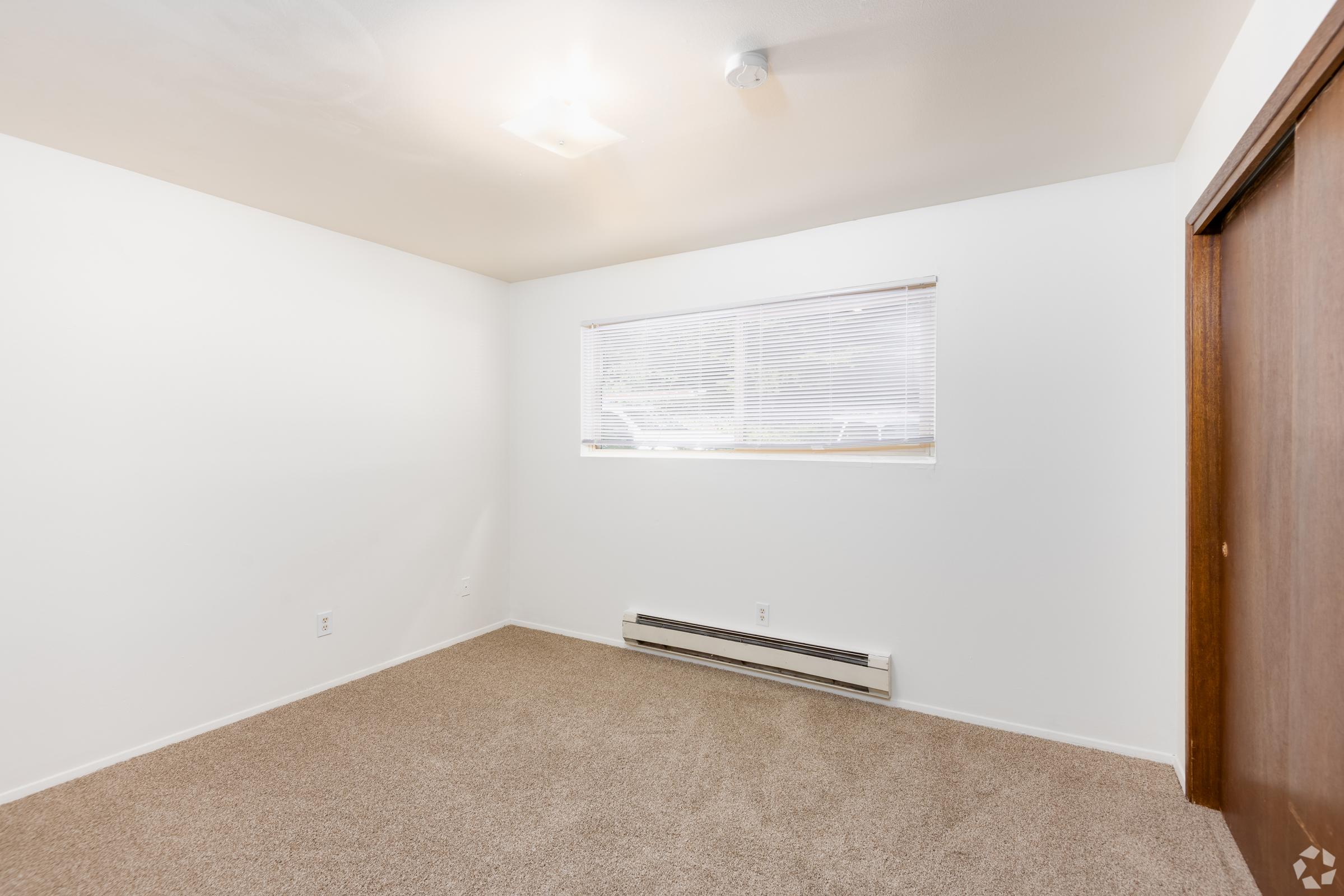 A well-lit empty room with light beige carpet and white walls. A small window with blinds allows natural light to enter, enhancing the bright and airy feel. The room features a heating unit along the wall and a wooden door. Ideal for use as a bedroom or office space.