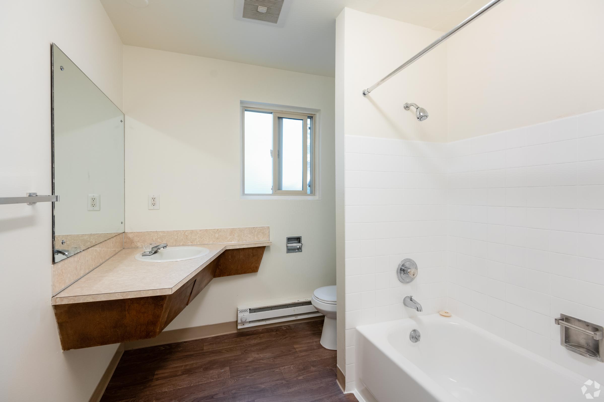 A clean and modern bathroom featuring a bathtub with a shower, a wall-mounted sink with a wooden shelf, and a large mirror. A small window allows natural light, and the floor is finished with dark wood. The walls are painted light colors, creating a bright and spacious feel.