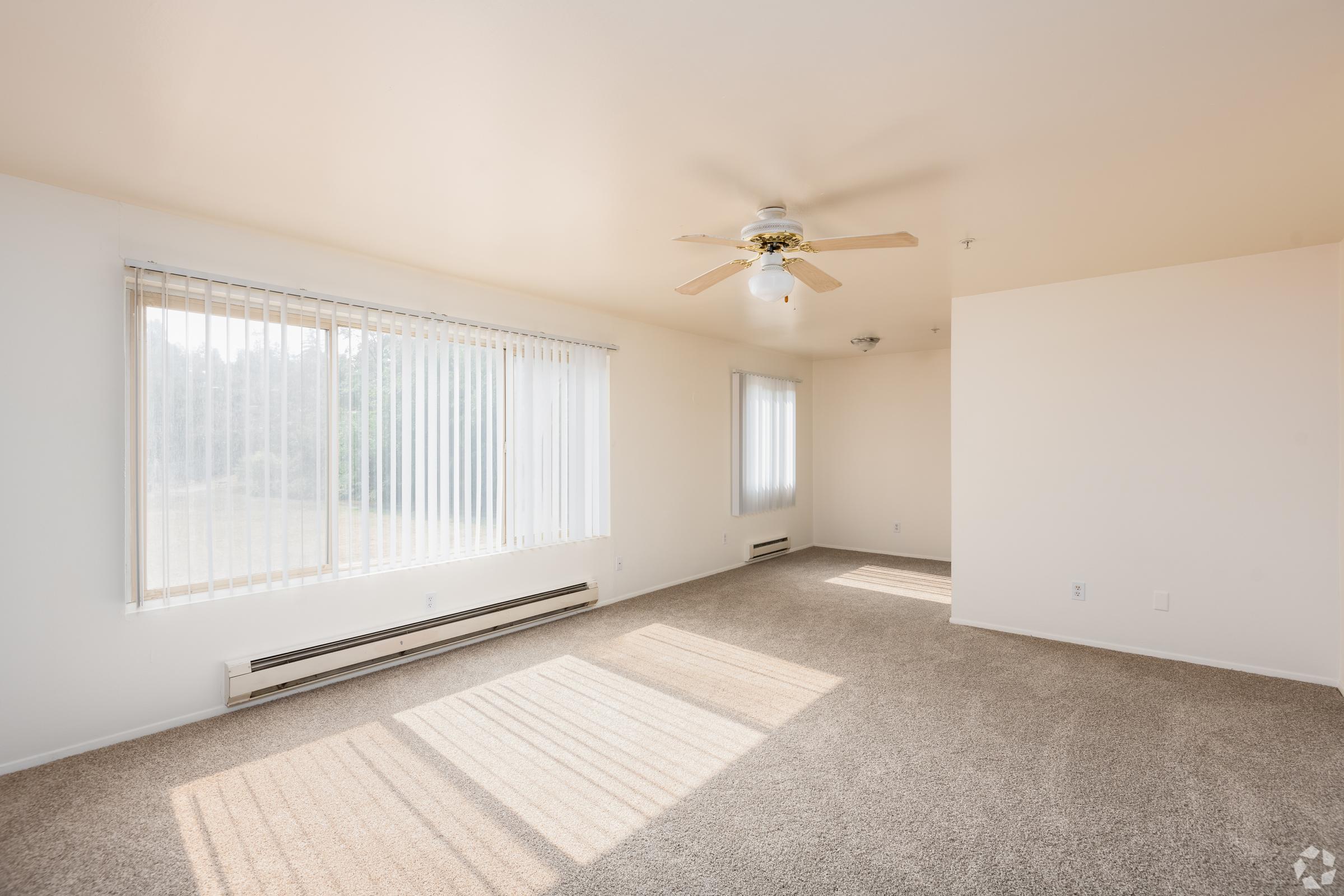 Bright and airy living room with beige carpet, white walls, and large windows allowing natural light. A ceiling fan with light fixture hangs from the ceiling, and there are heating vents along the walls. The room is empty and spacious, featuring a panoramic view of greenery outside.