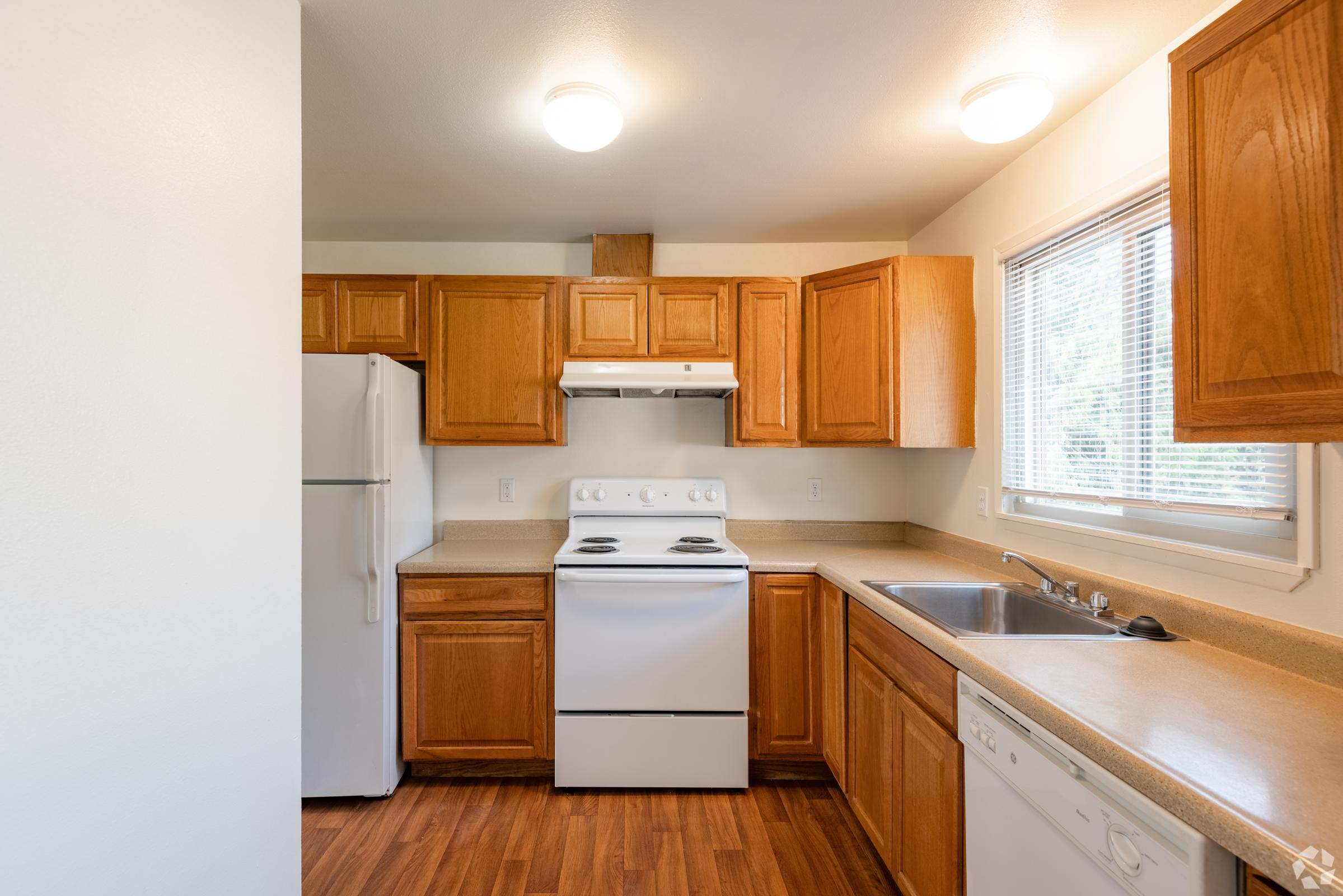 A modern kitchen featuring wooden cabinetry, a white stove and refrigerator, a sink with a window above, and light hardwood flooring. The space is well-lit with two overhead lights and has a clean, organized appearance.