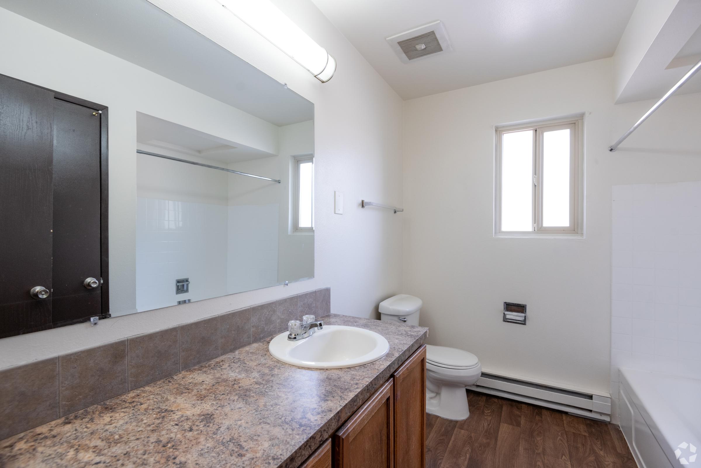 A modern bathroom featuring a sink with a brown countertop, a mirror above it, a white toilet, and a bathtub. Natural light enters through a small window. The walls are painted a light color, and there is a dark cabinet on the left side for storage.
