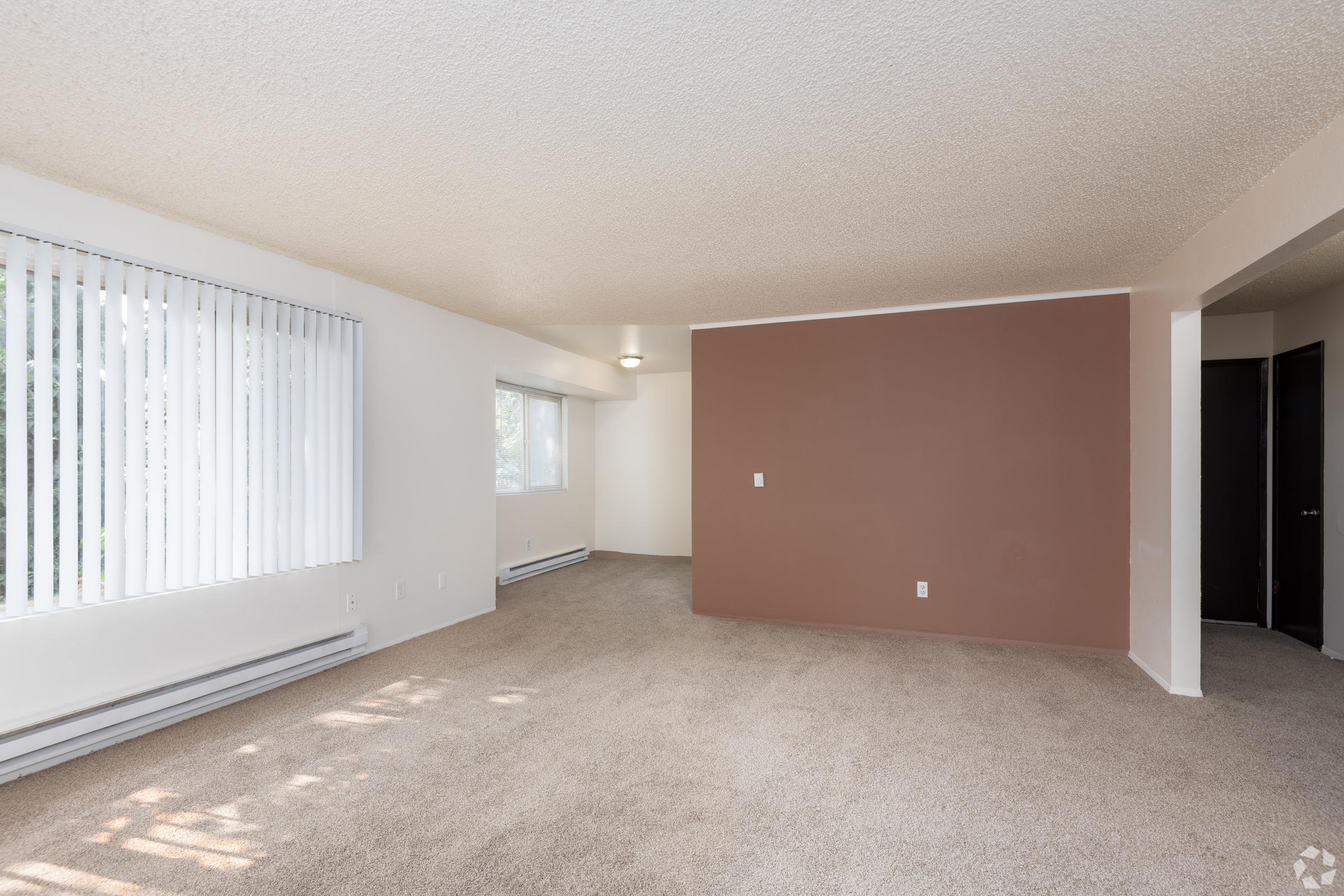 Spacious room with beige carpet, light-colored walls, and a brown accent wall. Large windows with vertical blinds allow natural light. Features include a carpeted area, baseboard heating, and a doorway leading to another room. The overall ambiance is bright and inviting.
