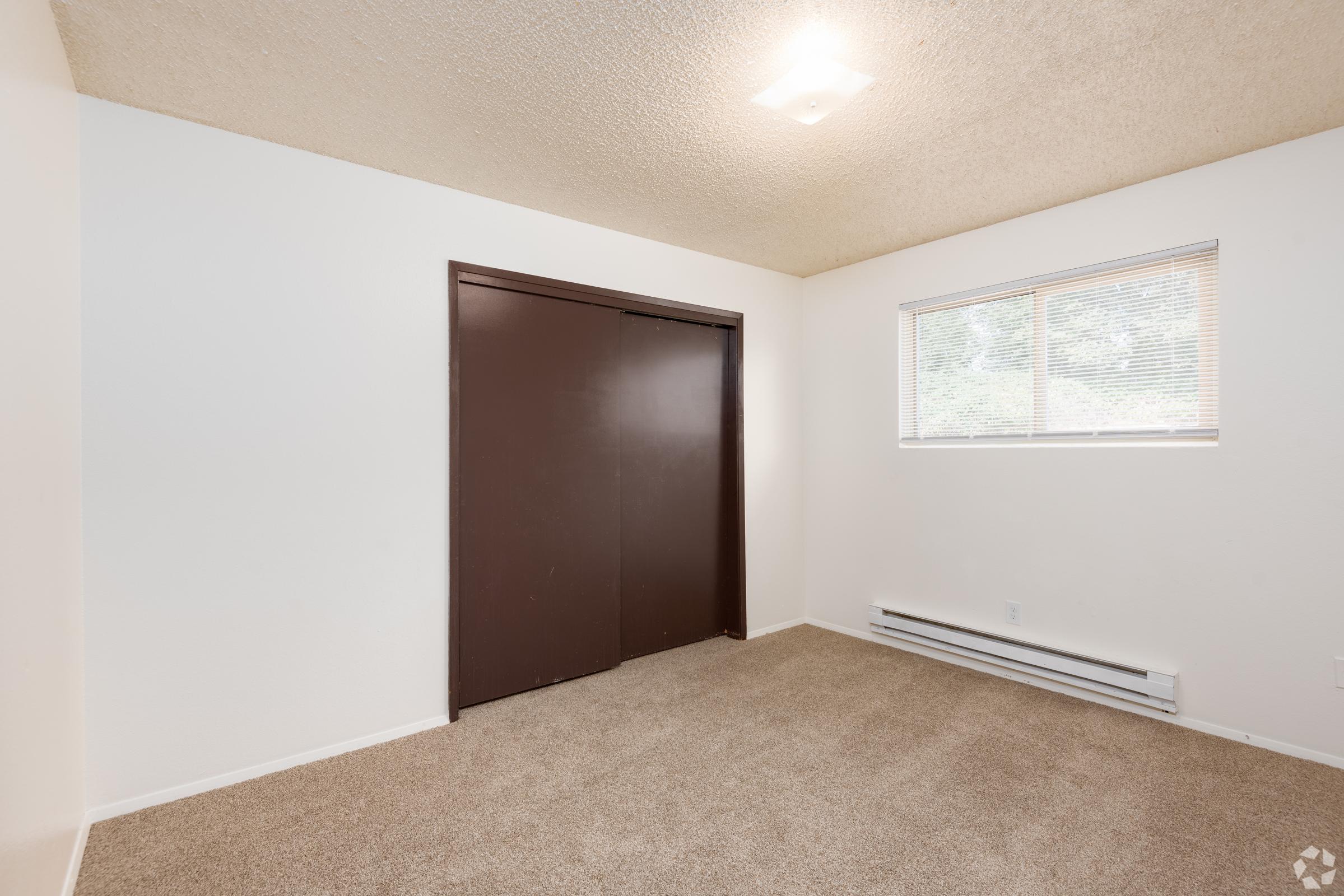 A vacant room featuring tan carpet, white walls, and a textured ceiling. There is a large brown closet door on the left side and a window on the right with blinds, allowing natural light to enter. The room is empty, suggesting it is ready for new furnishings.