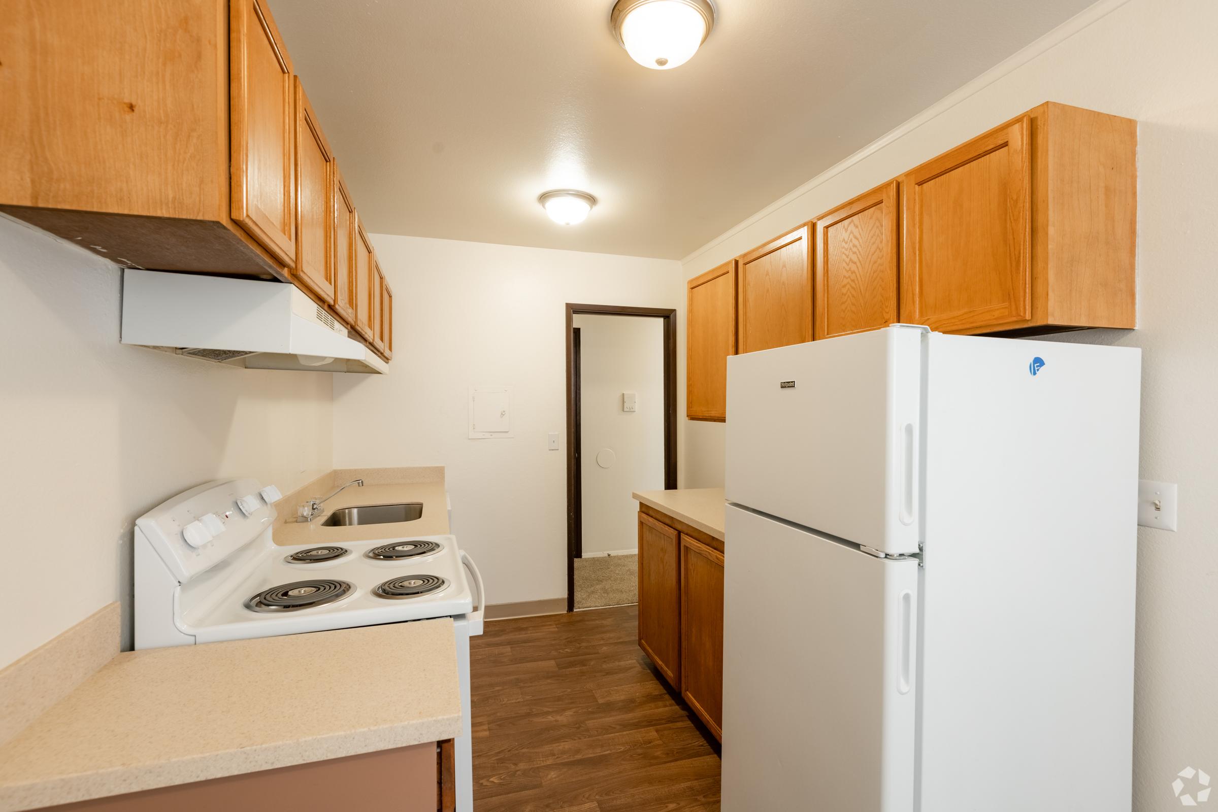 A small kitchen featuring wooden cabinets, a white refrigerator, a white stove with four burners, and a countertop. The walls are painted light, and there is a ceiling light fixture. The room appears well-lit and includes a doorway leading to another area.