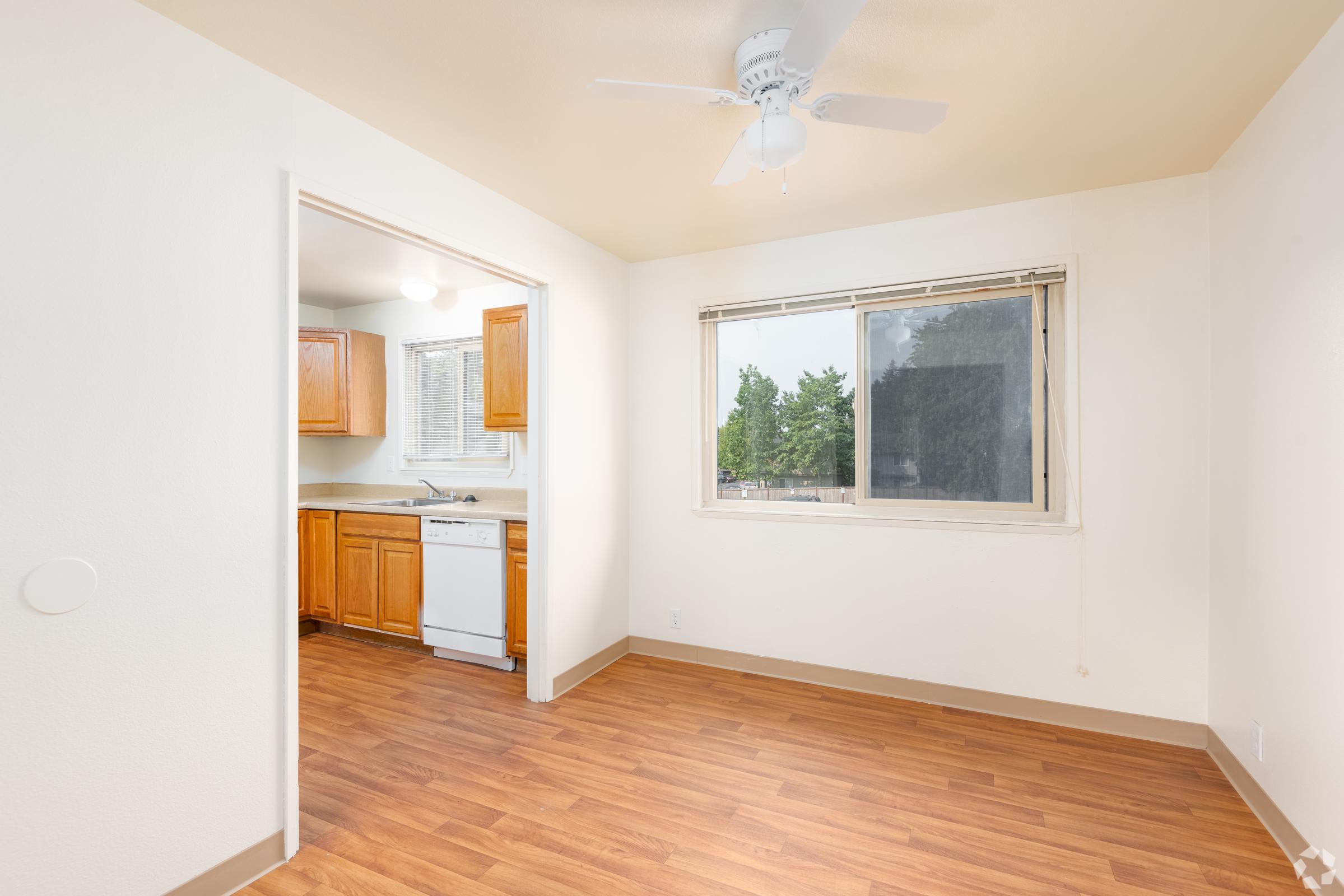 A well-lit, unfurnished room with wooden flooring, featuring an open doorway leading to a kitchen area with wooden cabinets. A window provides natural light, and a ceiling fan is visible. The walls are painted white, giving a bright and clean atmosphere.