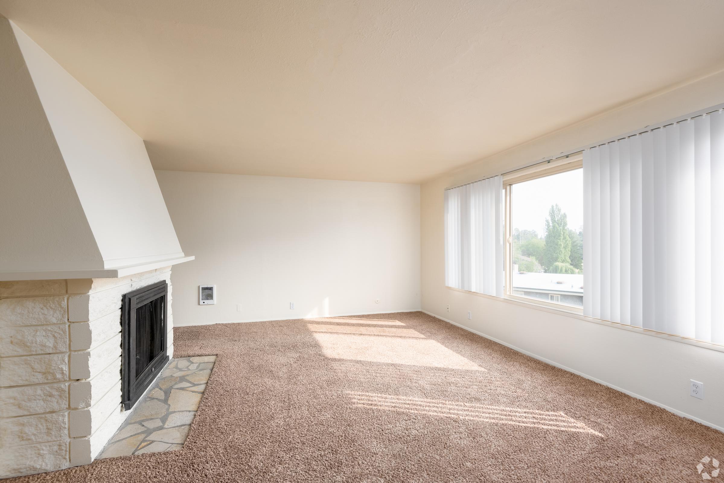 A spacious living room featuring a white stone fireplace, beige walls, and large windows with white vertical blinds. The floor is covered with light brown carpet, and natural light streams in, highlighting the room's emptiness and neutral aesthetics.