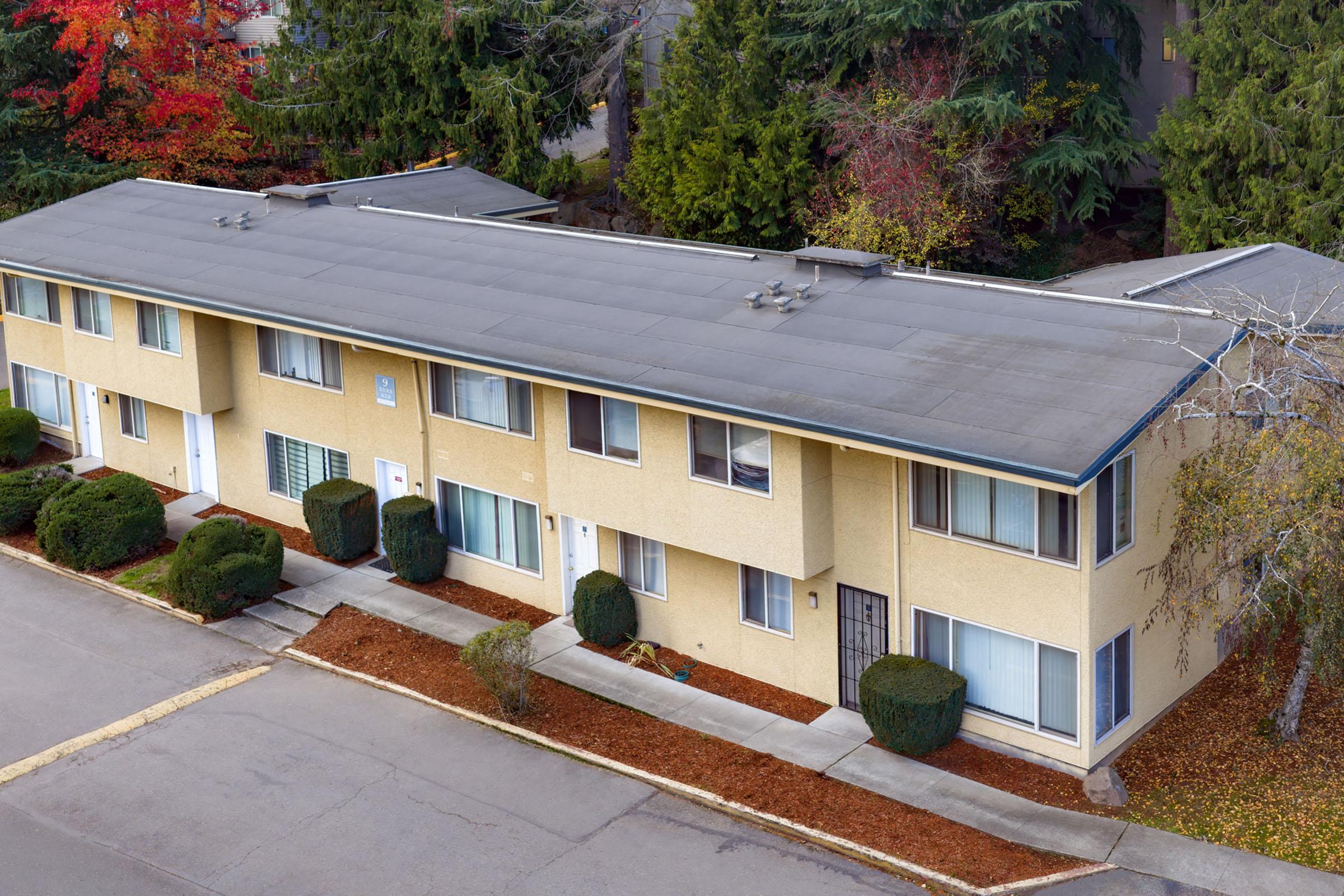 Aerial view of a single-story apartment building with a flat roof, surrounded by manicured bushes and a tree-lined street. The building features several windows and a few entry doors. Autumn foliage adds color to the scene.