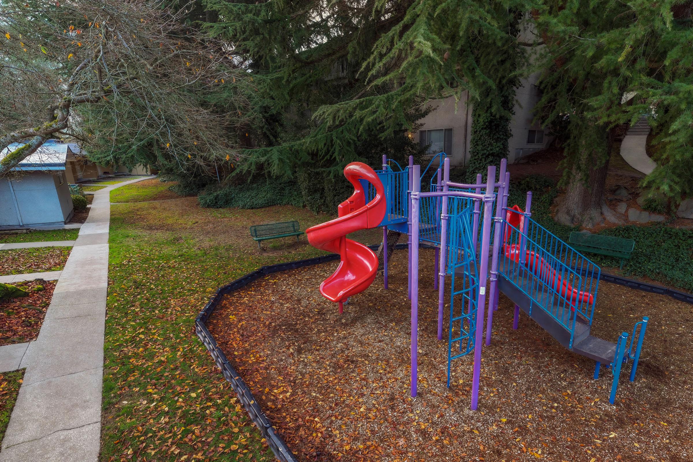 A colorful playground featuring a red slide and climbing structure, surrounded by green grass and trees. A pathway can be seen nearby, along with benches for seating. The scene is tranquil and inviting, suitable for children to play and explore.
