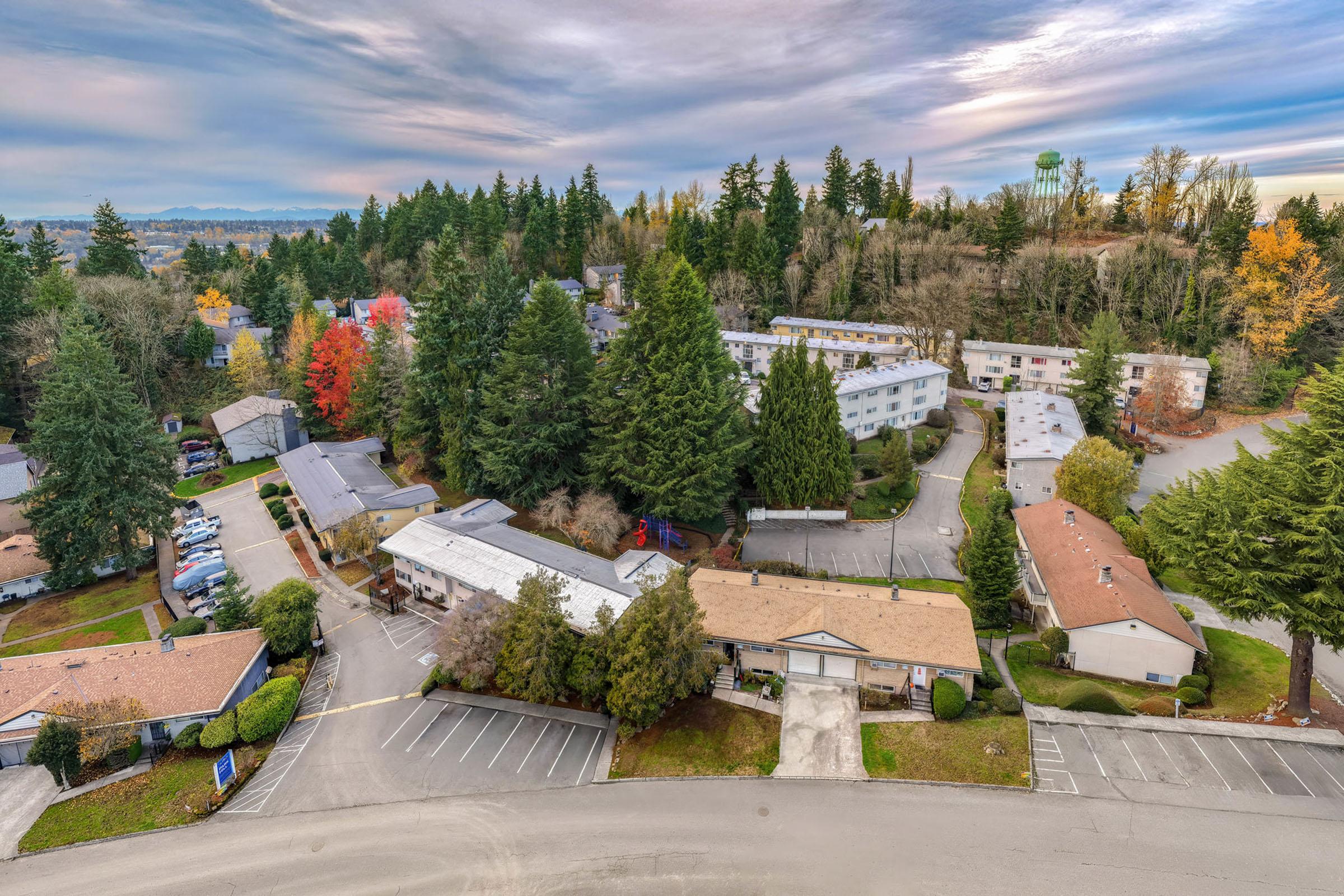 Aerial view of a residential area surrounded by lush trees, featuring several low-rise buildings with varying roof colors. The layout includes parked cars and pathways, with a backdrop of hills and a cloudy sky, showcasing the tranquility of suburban living.
