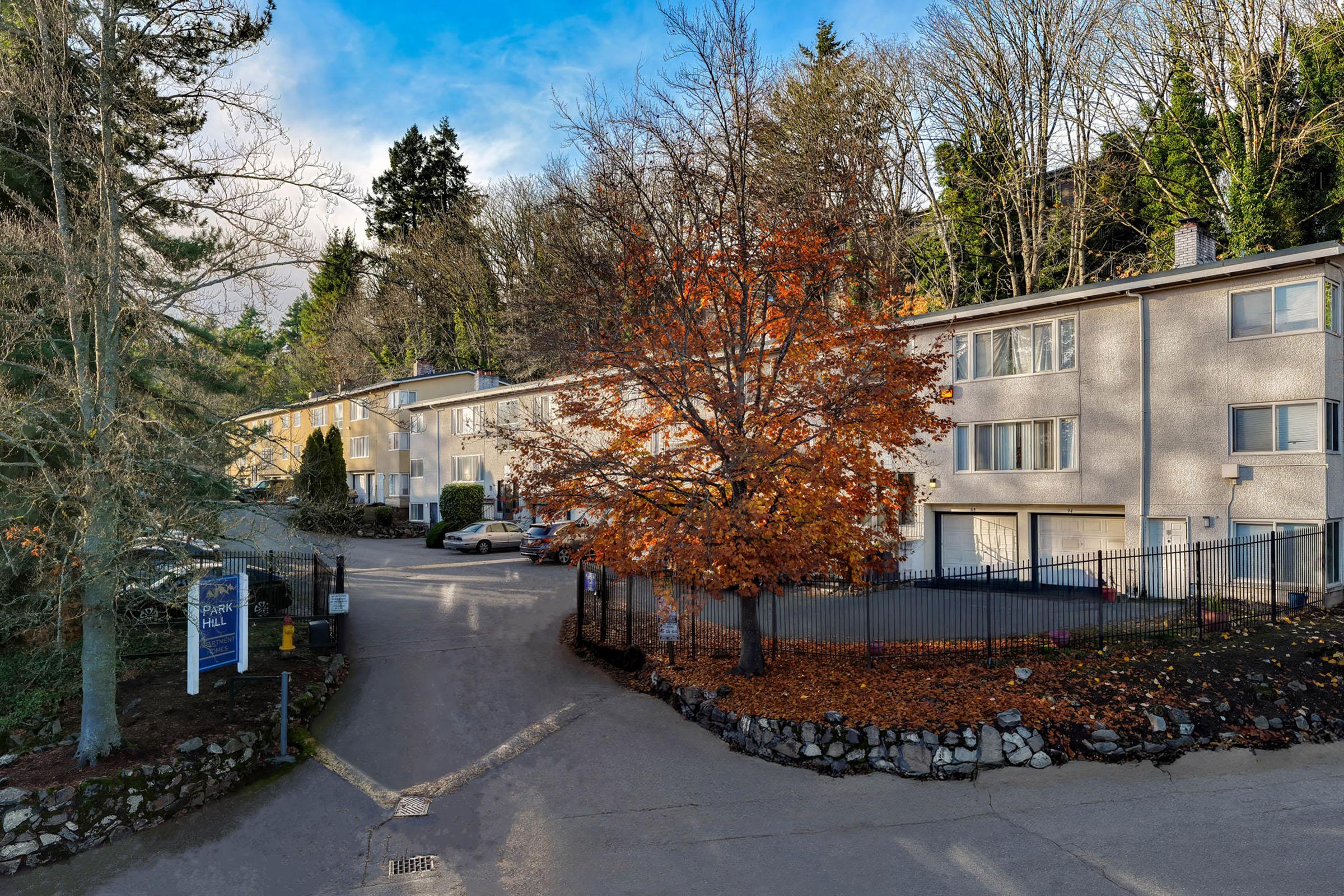 A residential area featuring low-rise apartment buildings surrounded by trees. A large tree with vibrant orange leaves stands in the foreground, and a driveway leads into the complex. A peaceful atmosphere is captured with clear skies and greenery in the background.