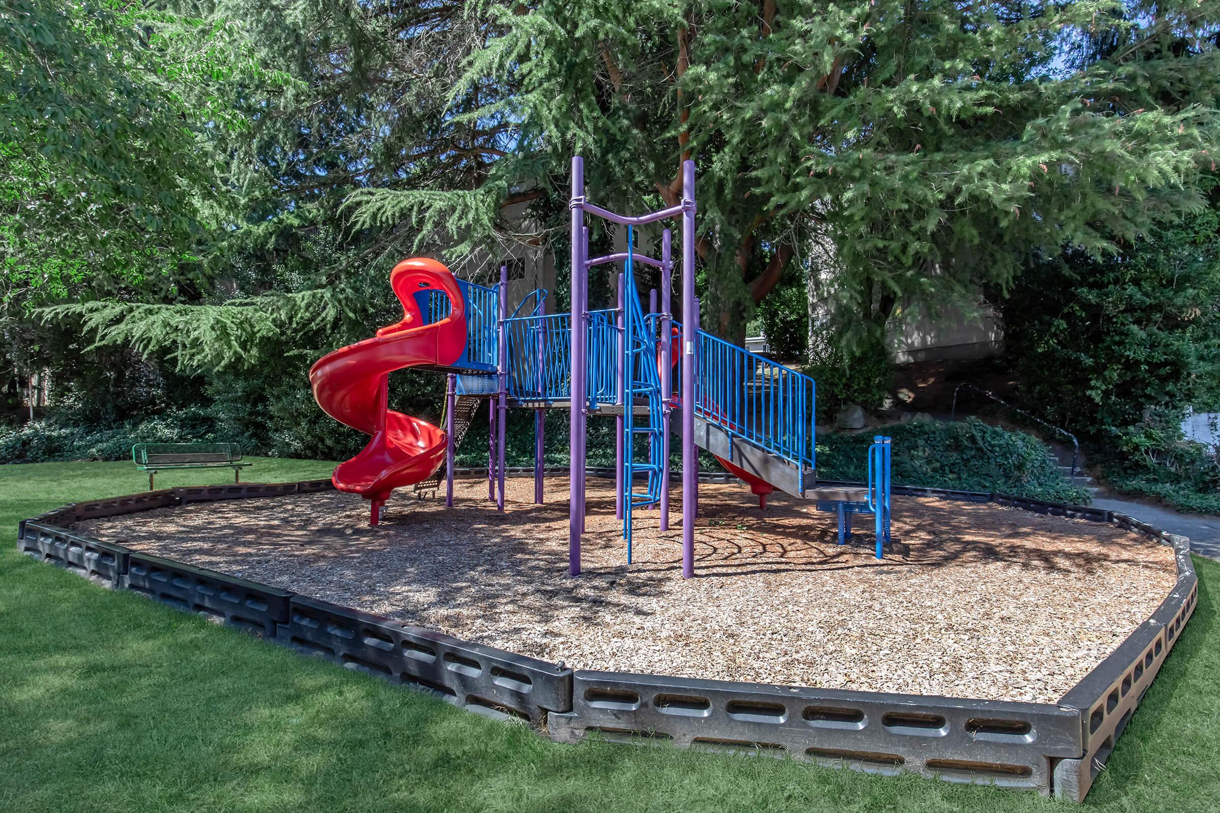 Colorful playground structure featuring a red slide, climbing area, and various play equipment, surrounded by a grassy area and trees, with a gentle wood chip surface beneath.