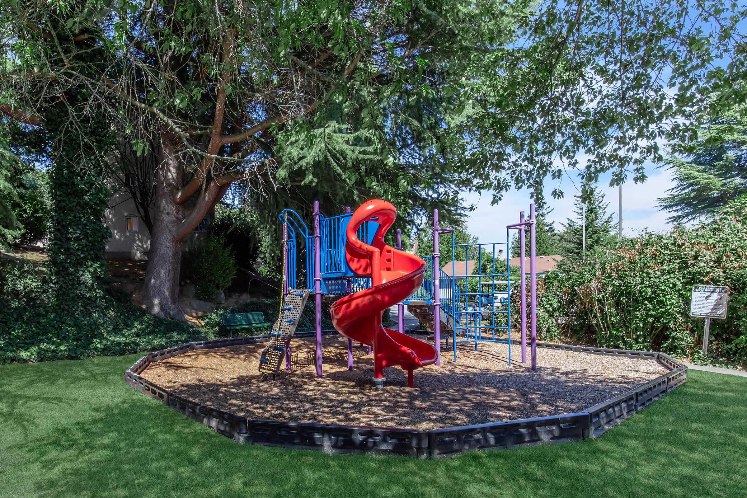 A playground featuring a bright red slide and colorful climbing structures, surrounded by lush green trees. The area has a soft, grassy surface and is designed for children's play, providing a safe and inviting environment. Sunny weather enhances the cheerful atmosphere.