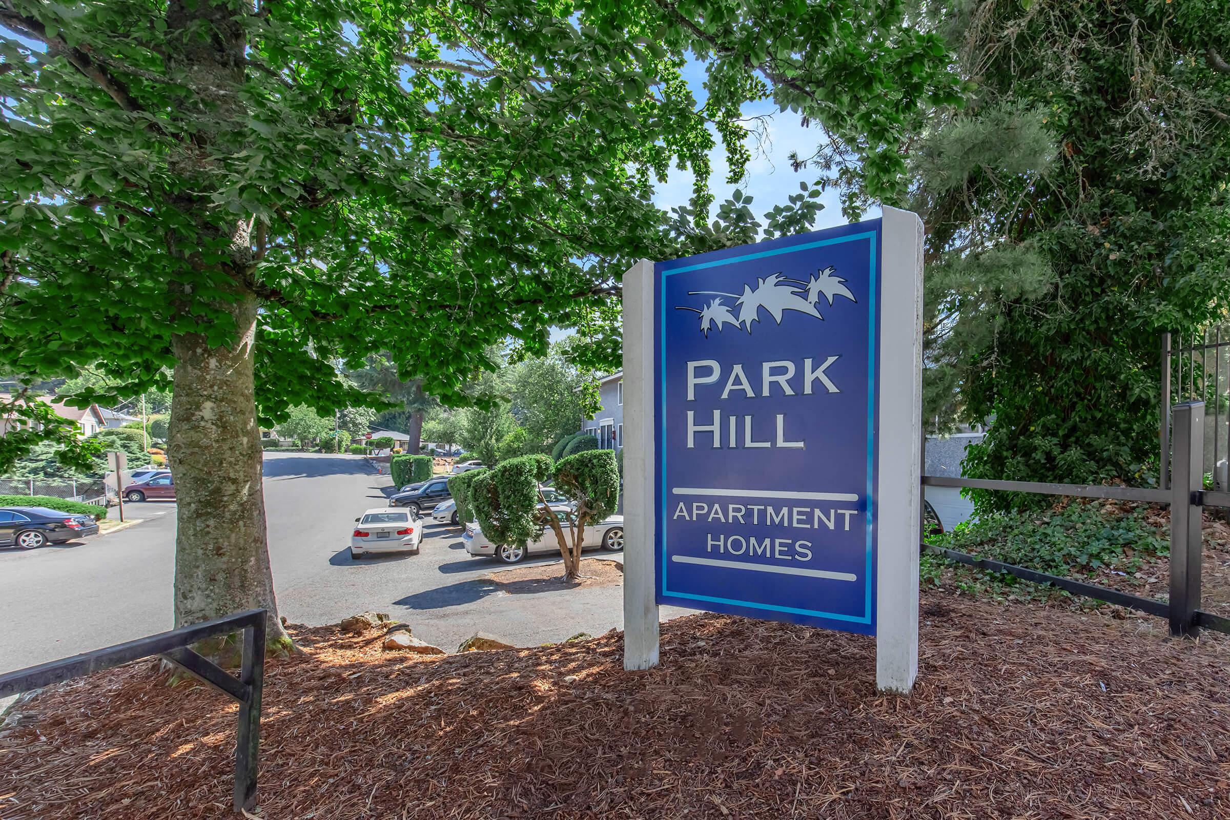 Sign for Park Hill Apartment Homes, featuring a blue background with white lettering and palm tree graphics. The sign is surrounded by lush green trees and landscaping, with parked cars visible in the background on a well-maintained road.