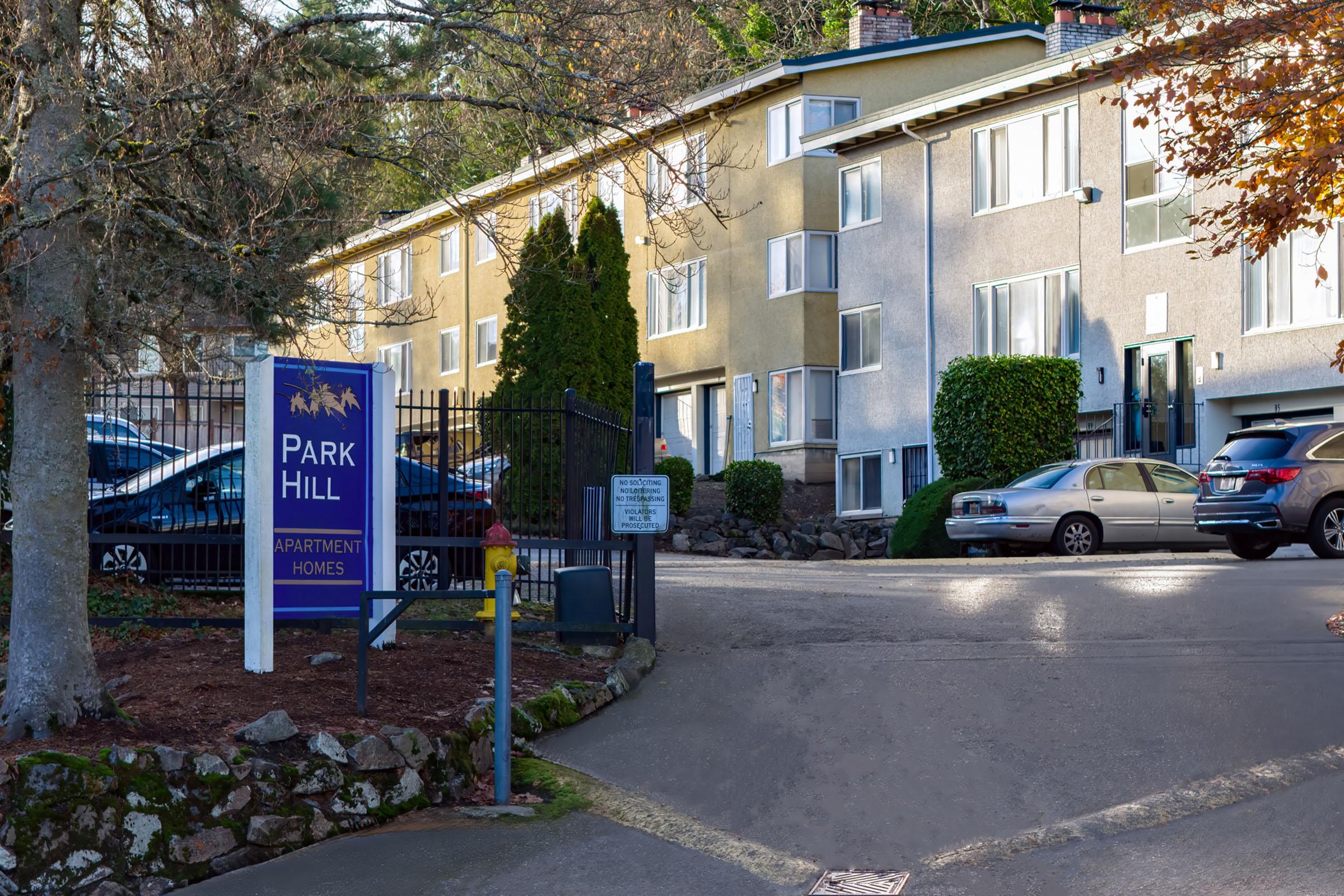 Entrance to Park Hill apartment homes featuring a sign, well-maintained landscaping, and a parking area. Three apartments can be seen with large windows and a gated entrance lined with trees.
