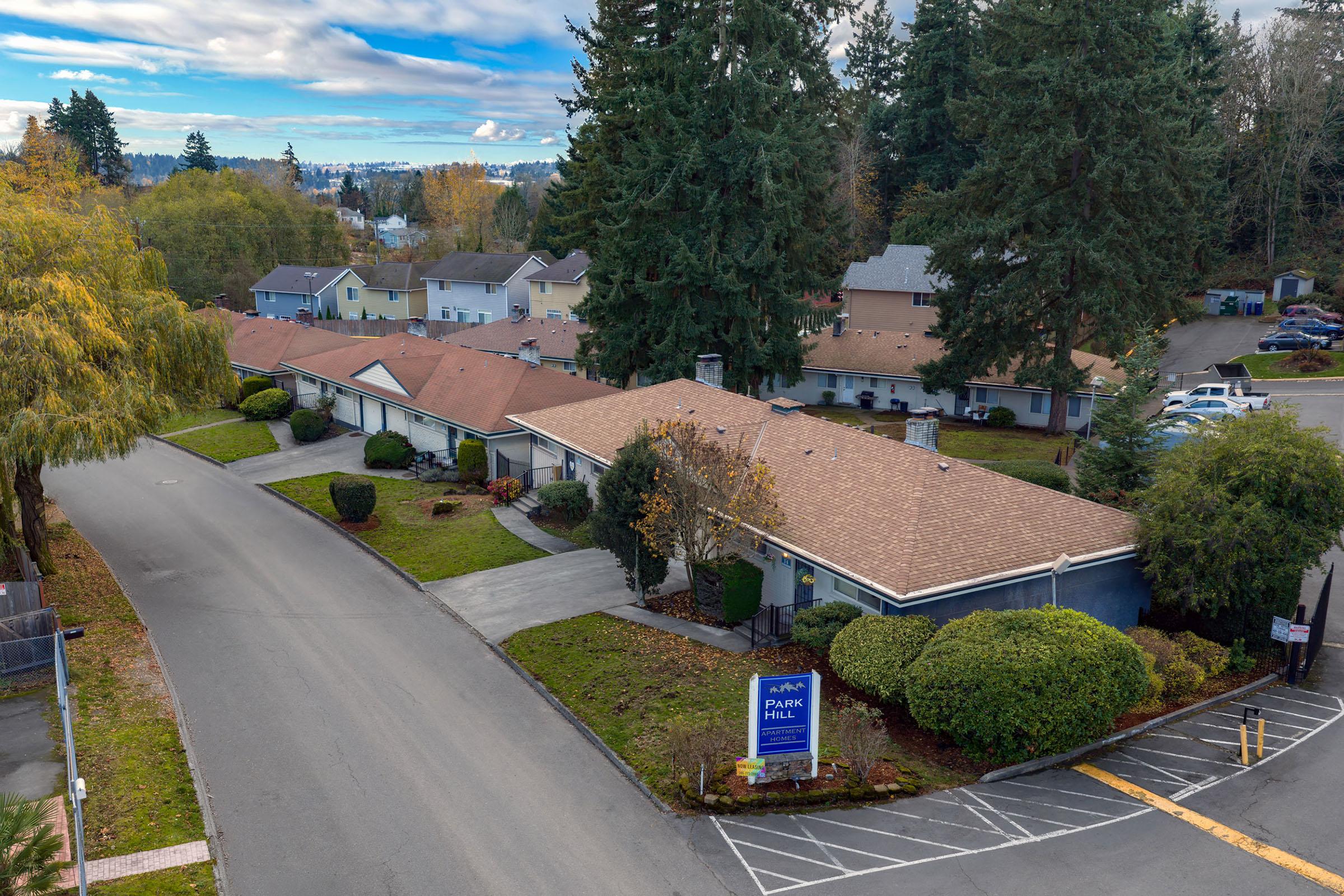 Aerial view of a residential community featuring single-story buildings with brown roofs, landscaped gardens, and a circular driveway. The sign for "Alpha Park" is visible at the entrance. Surrounding trees and autumn foliage create a scenic atmosphere.