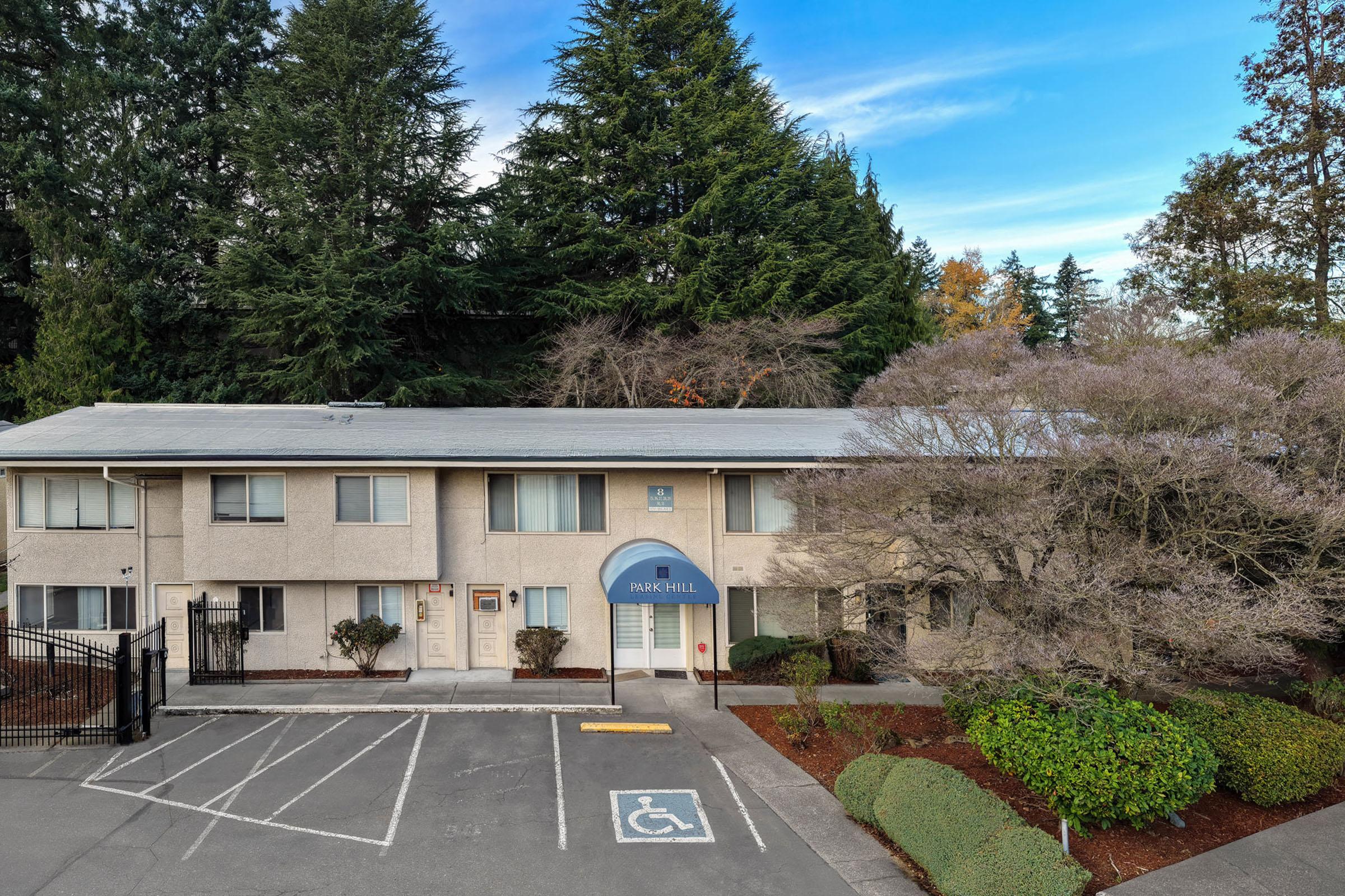 A view of a residential building named "Parkhill" surrounded by trees, with a landscaped parking area that includes a designated handicapped space. The building features a gray roof, light-colored exterior, and is set against a backdrop of green foliage and a clear blue sky.