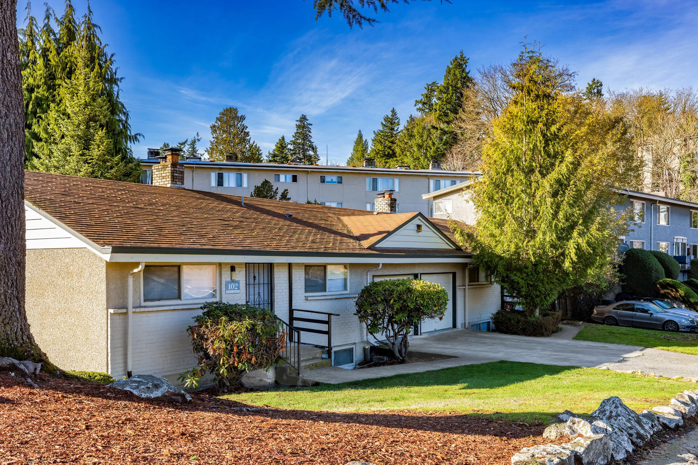 A residential house with a sloped roof, surrounded by trees and shrubs. The driveway is visible in front of the house, and there are other homes in the background under a clear blue sky. The setting is peaceful and suburban, with green foliage nearby.