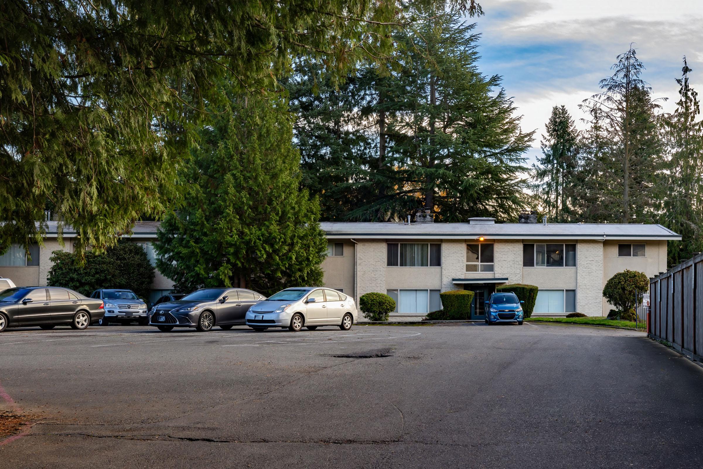 A residential building with a row of parked cars in front. The structure features a beige exterior and green shrubbery around it. Tall trees are visible nearby, and the sky is partly cloudy, creating a calm suburban atmosphere. The pavement shows some wear and tear.
