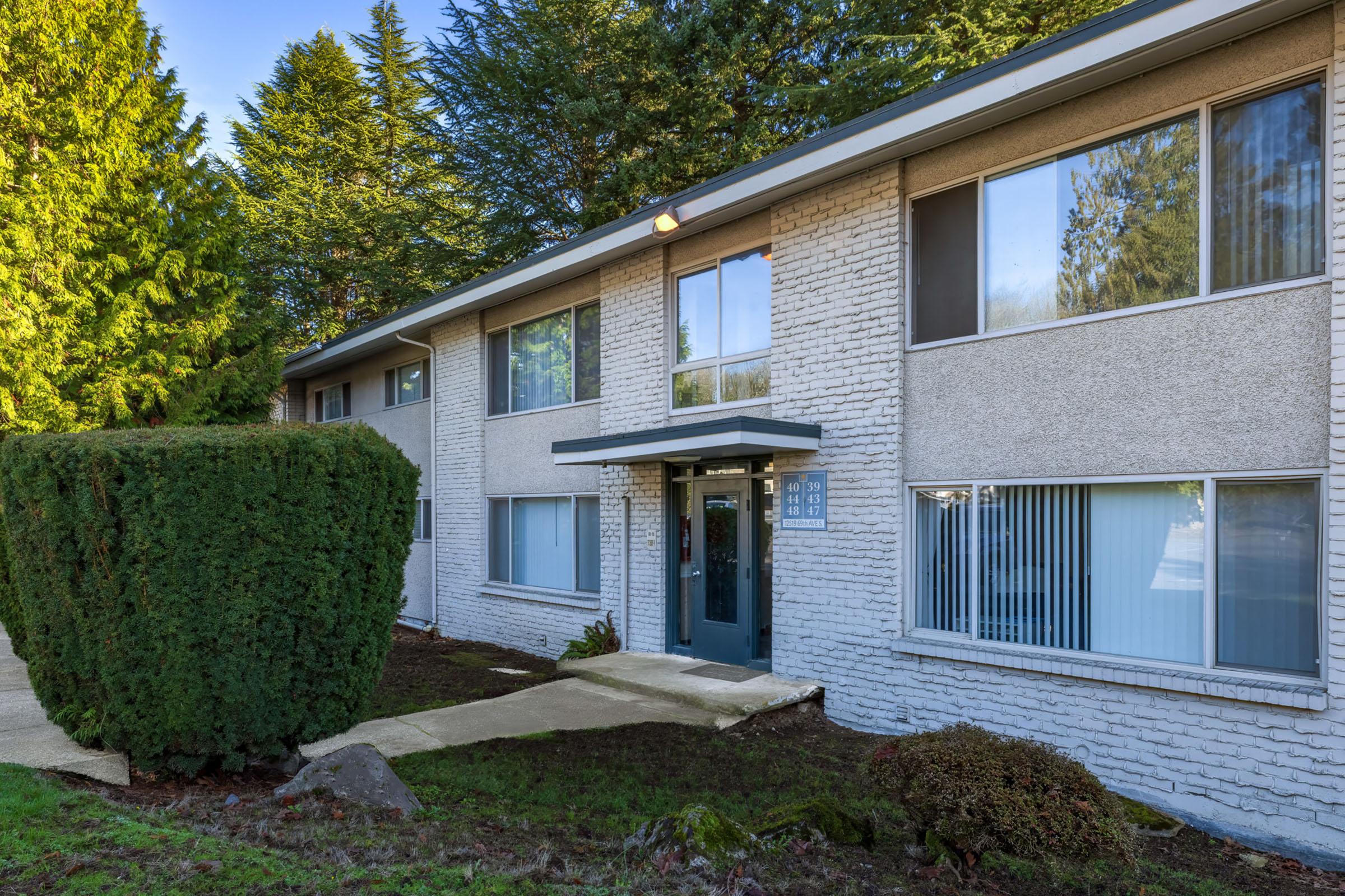 A modern, two-story apartment building with a gray exterior. The entrance features large windows and a doorway framed by greenery. A neatly trimmed shrub is visible in front, and the lawn is well-maintained, with trees in the background suggesting a peaceful residential setting.