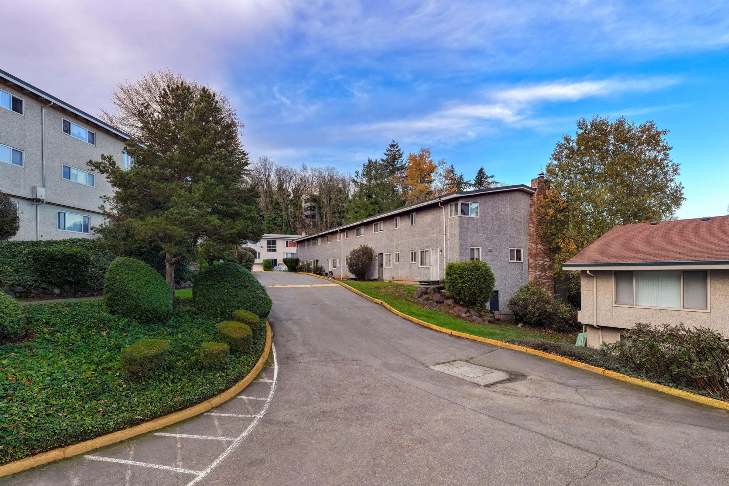 A residential complex featuring multiple buildings set among lush greenery. The scene includes a curving driveway lined with neatly trimmed bushes and trees. The sky is partly cloudy, adding a serene ambiance to the environment.