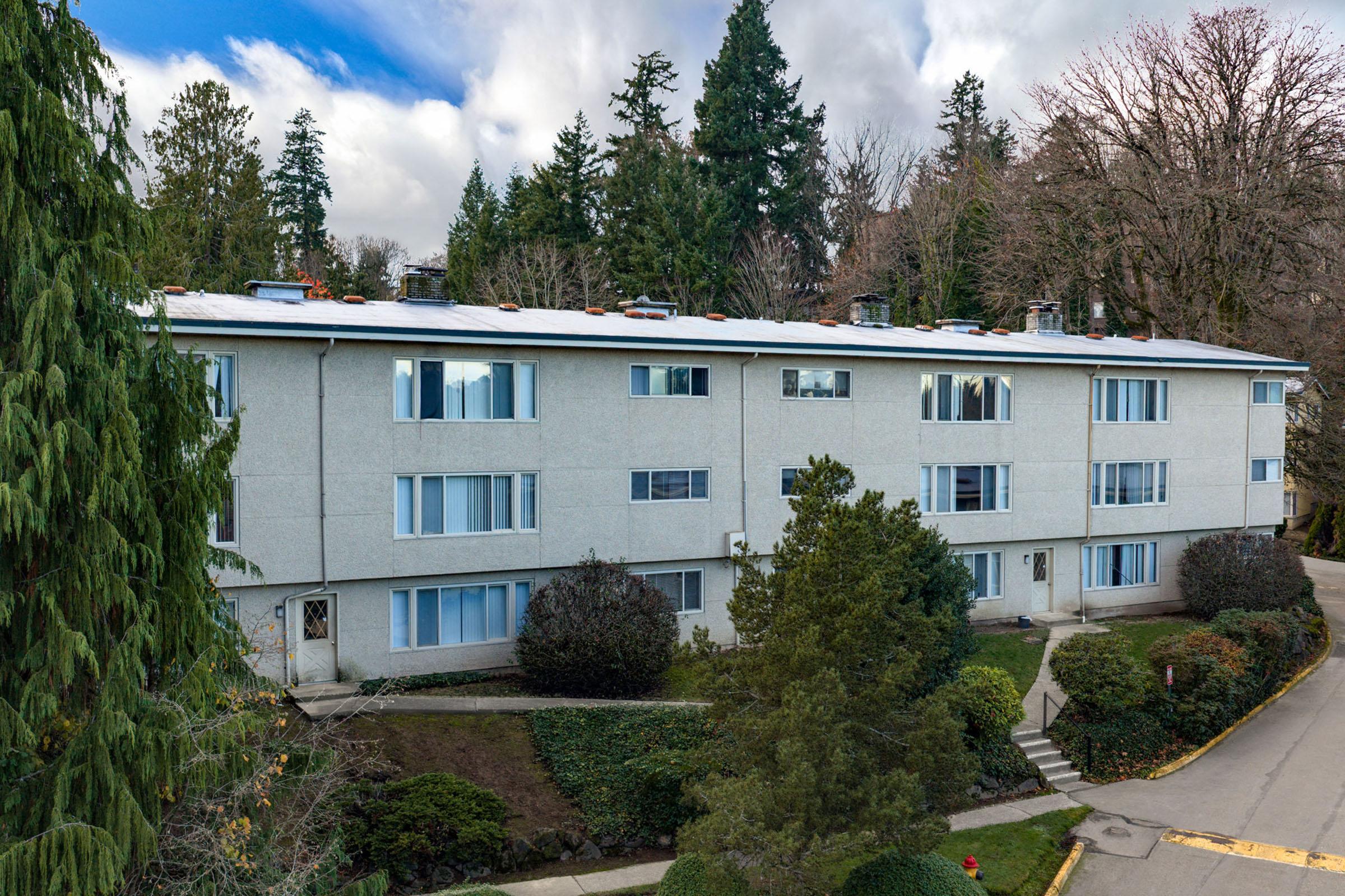A light grey, multi-unit apartment building with rows of windows on the front, surrounded by well-maintained shrubs and trees. The sky is partly cloudy, and the scene evokes a tranquil suburban atmosphere. The building features a flat roof and a paved pathway leading to the entrance.