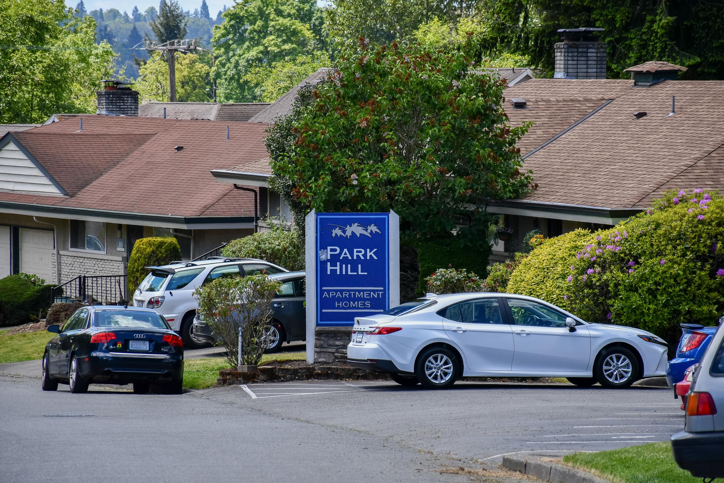 Sign for "Park Hill Apartment Homes" in front of a residential area with several parked cars and greenery. The buildings have brown roofs and the surroundings include trees and shrubs. The scene is set in a calm, suburban environment.
