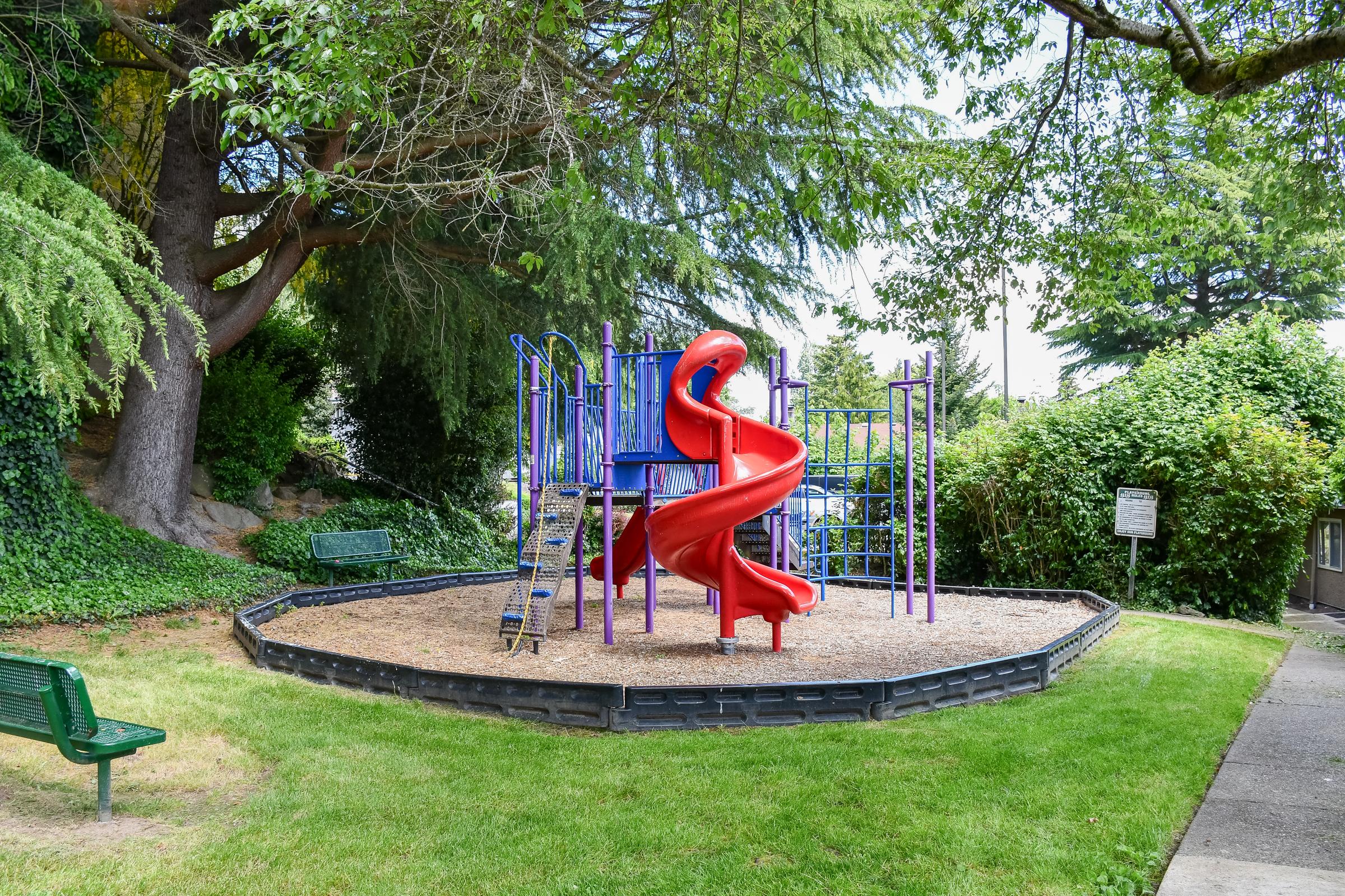 A colorful playground featuring a red slide, purple climbing structures, and a sandy base. Surrounded by lush greenery and trees, with a park bench nearby. Ideal for children's outdoor play.