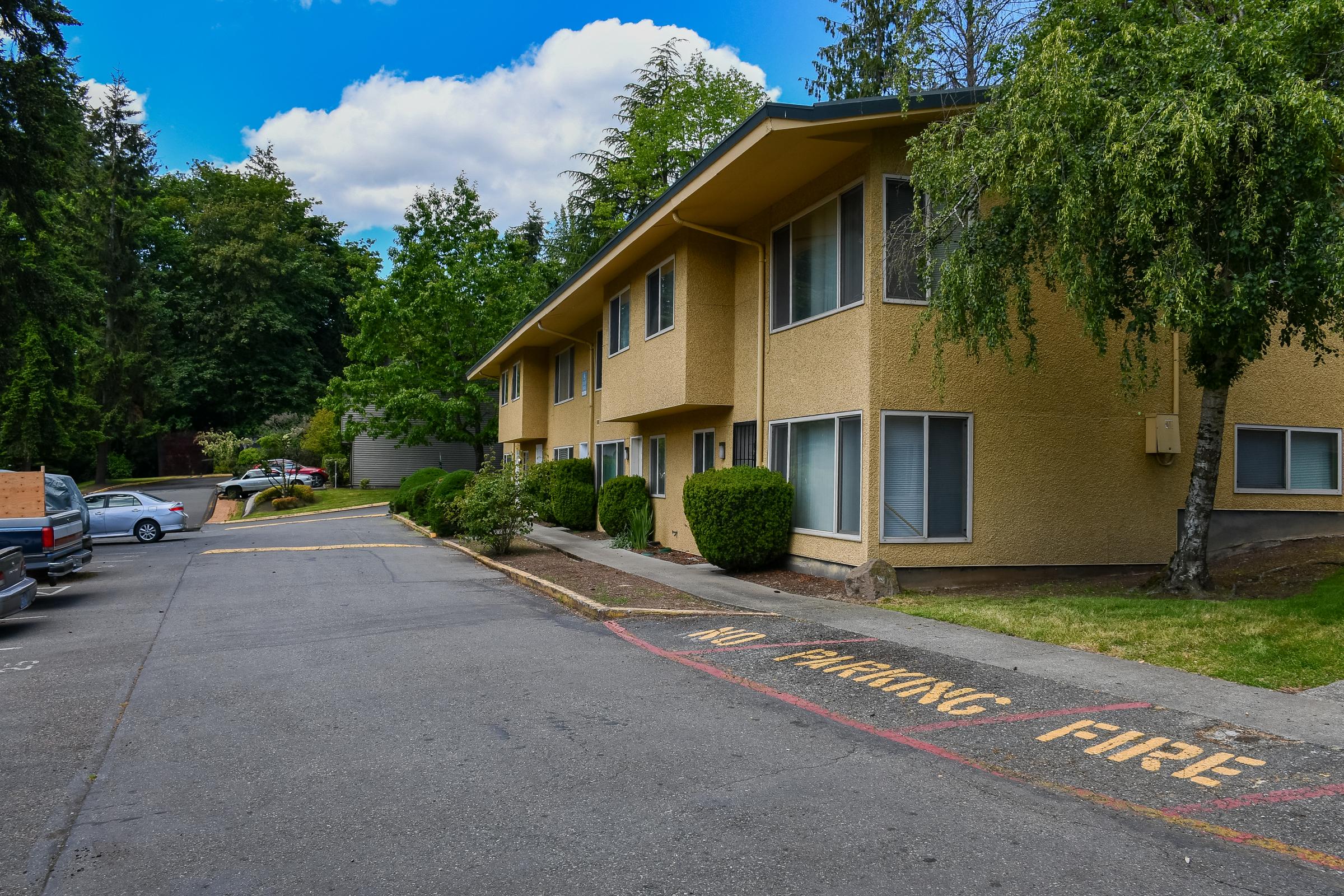 A view of an apartment building with yellow exterior walls, surrounded by trees and shrubs. In the foreground, a paved area with a "No Parking Fire" sign painted on the ground. The scene includes parked cars and a cloudy blue sky above.