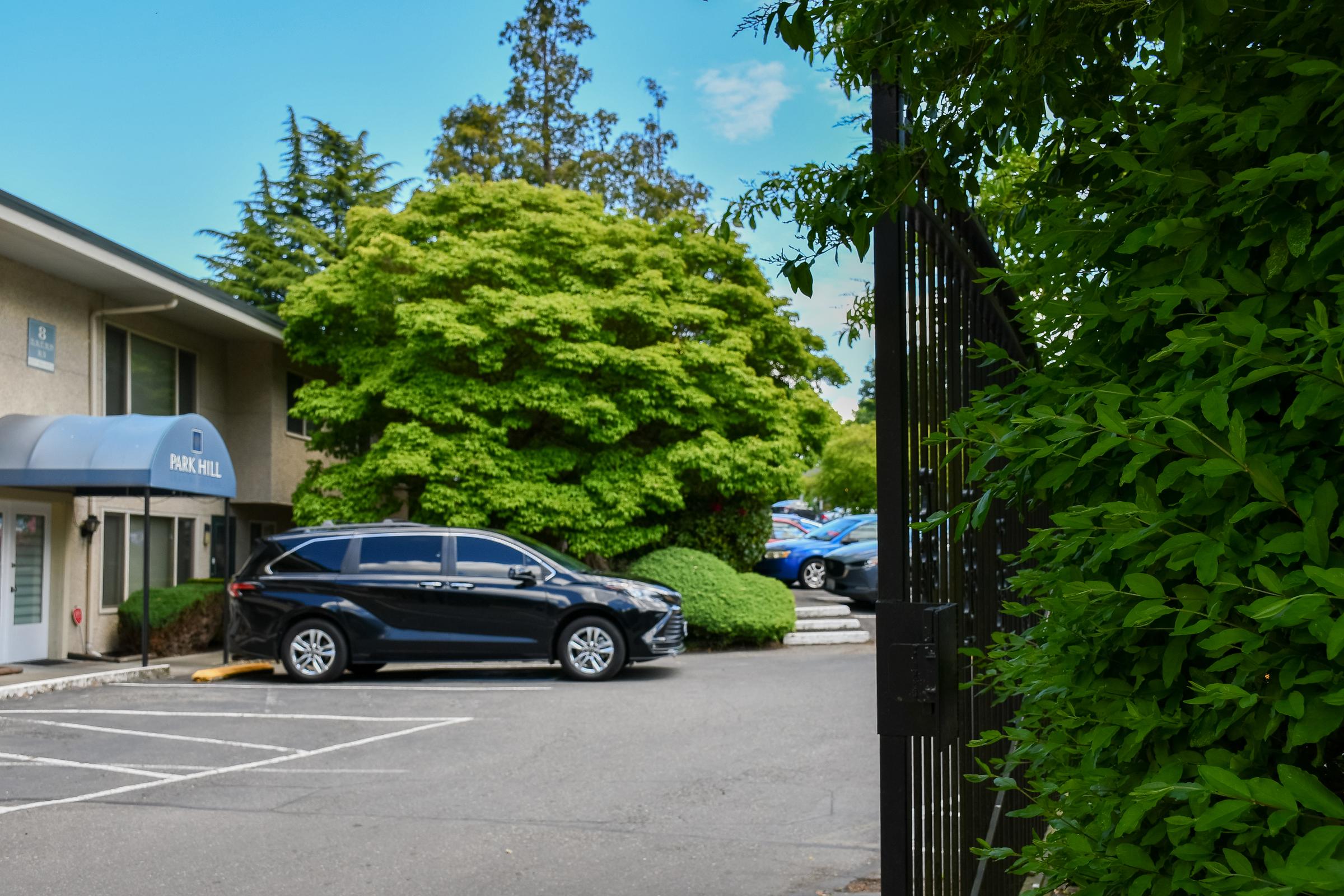 A black SUV parked in a parking lot next to a green shrub. In the background, trees and a building are visible, with a sign indicating "PARKING." The scene is set on a clear day with a blue sky. The gate on the right is partially open, leading into the parking area.