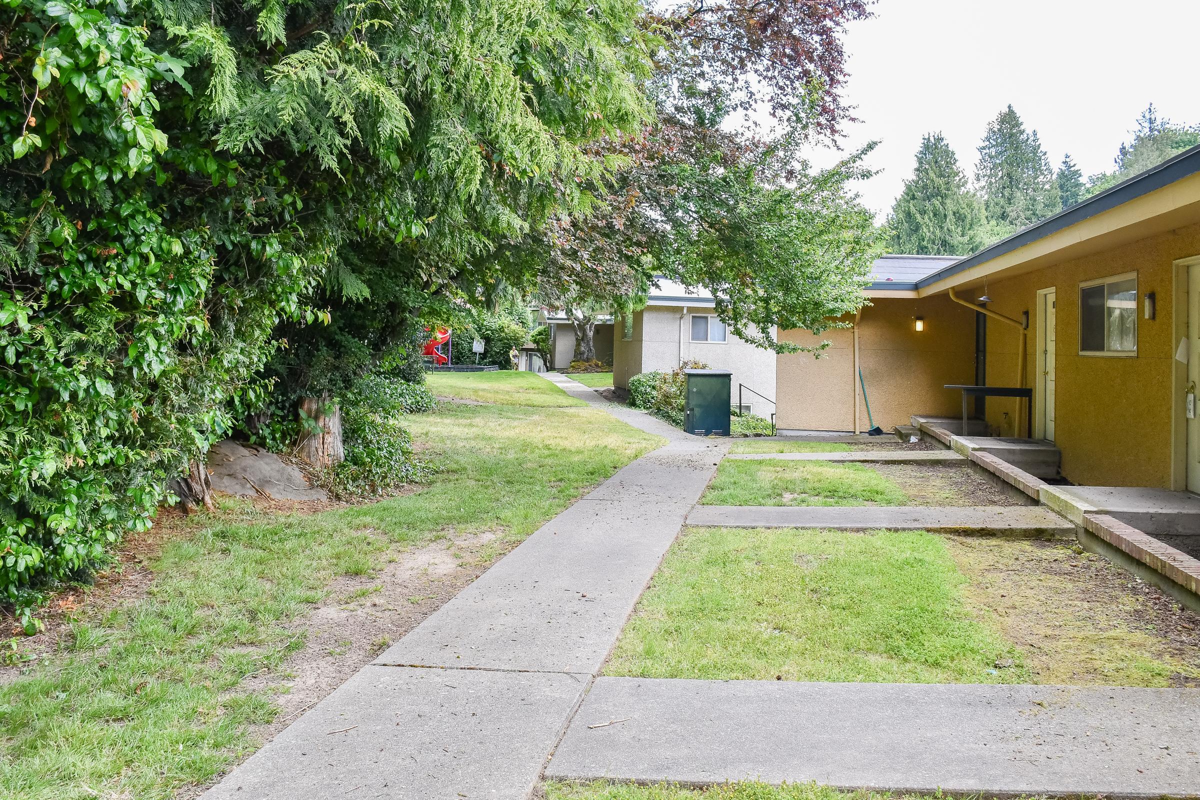 A pathway running through a grassy area leads to two buildings set among trees and shrubs. One building is yellow with a small porch, while the other is partially visible. The scene is peaceful, with greenery on one side and a small figure in the background.