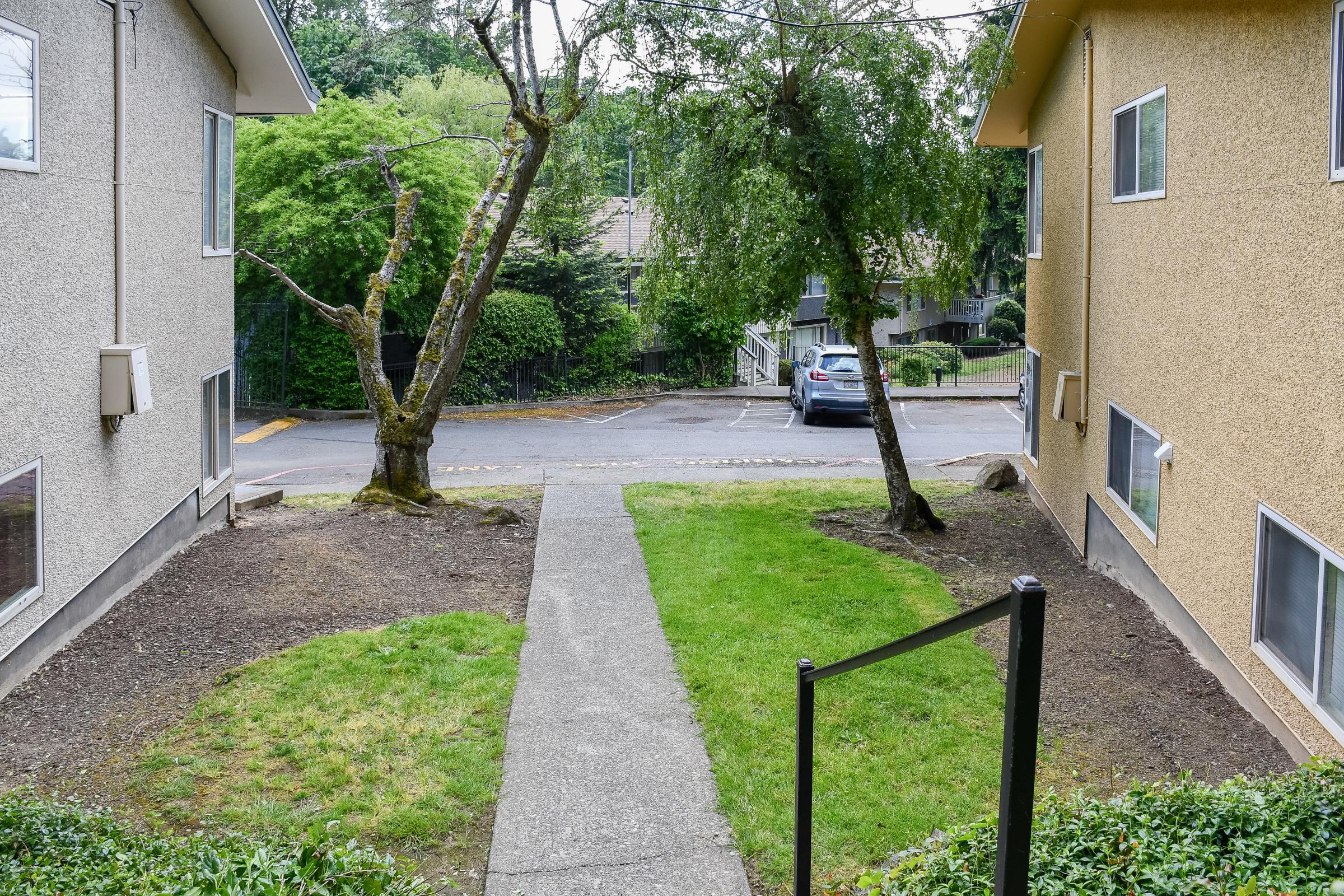 A view from a pathway between two apartment buildings, leading to a grassy area with trees on either side. A car is parked in the background, and the scene is surrounded by greenery, creating a calm residential atmosphere.