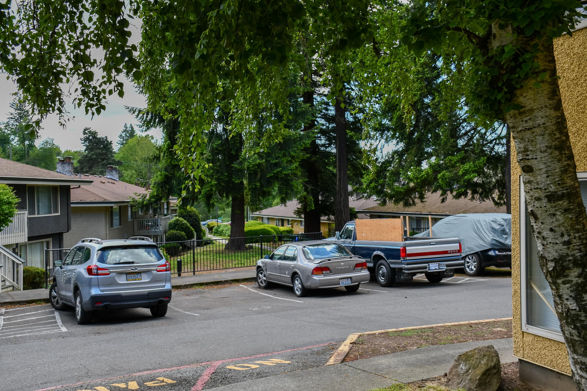 A view of an apartment parking lot with three vehicles parked, including a gray sedan and a pickup truck. There are trees in the background, a nearby fence, and several apartment buildings visible. The area appears well-maintained with green landscaping.