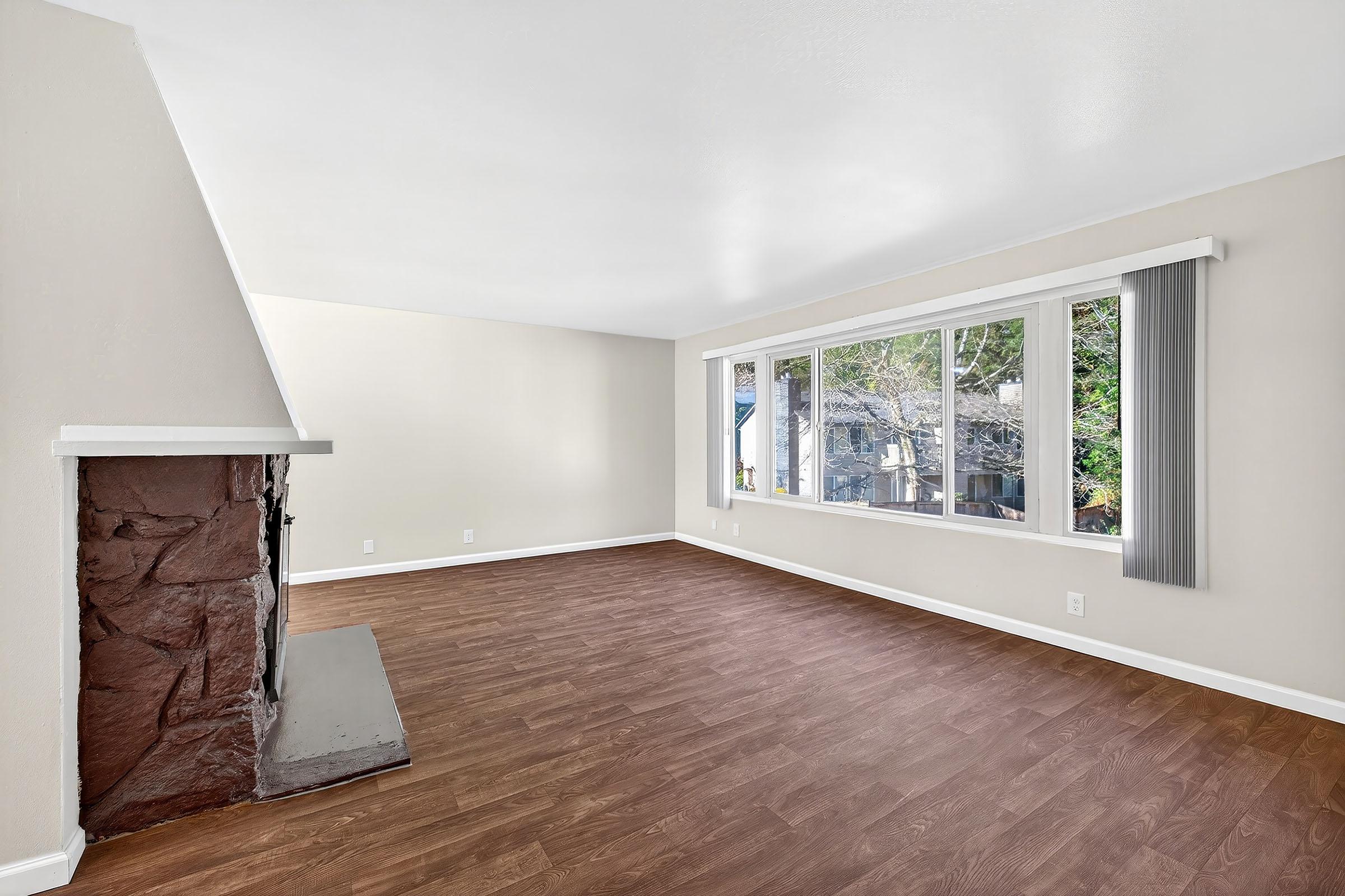 Spacious, well-lit interior of a living room featuring a stone fireplace on the left, large windows allowing natural light, light-colored walls, and a wooden floor. The room appears clean and ready for decoration, with a view of greenery outside through the windows.