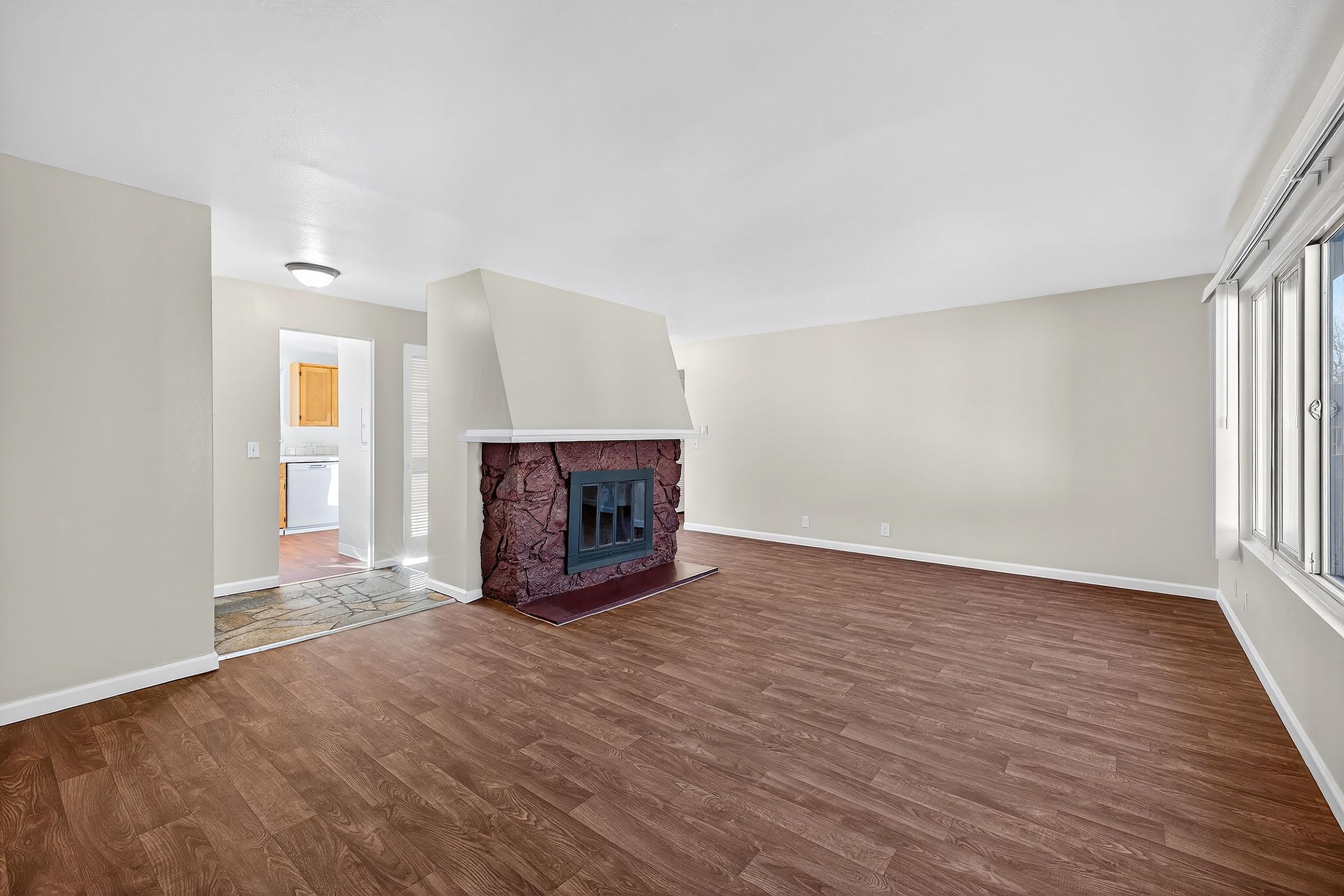 Empty living room with light-colored walls and a wooden floor. A stone fireplace is located on one wall, and there's a doorway leading to a kitchen area in the background. Large windows allow natural light, creating a spacious and airy atmosphere.