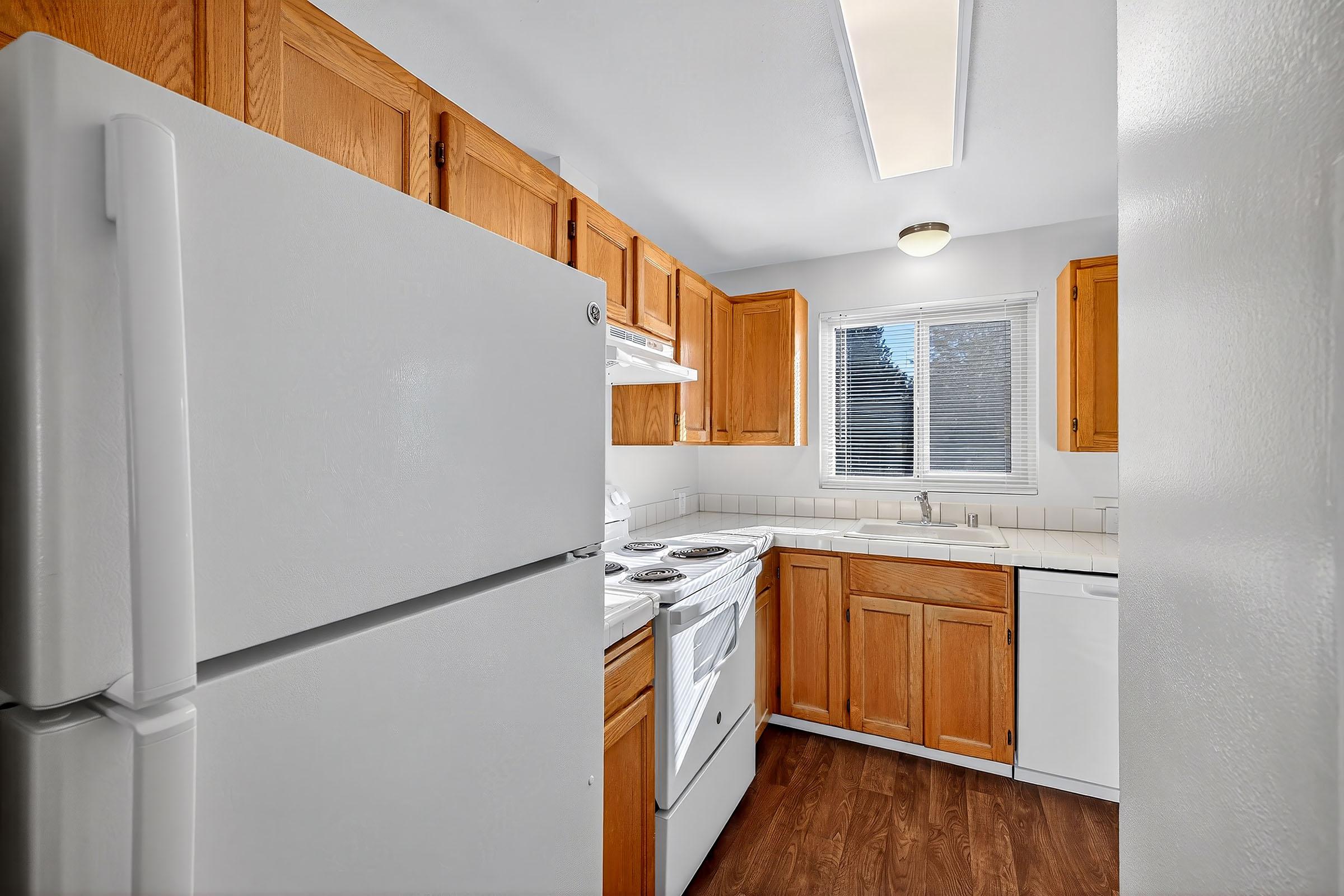 Bright kitchen with wooden cabinetry, a white refrigerator, and a stove. It features a sink below a window, allowing natural light to illuminate the space. The floor is made of dark wood, creating a warm atmosphere. The kitchen is clean and uncluttered, emphasizing functionality and comfort.