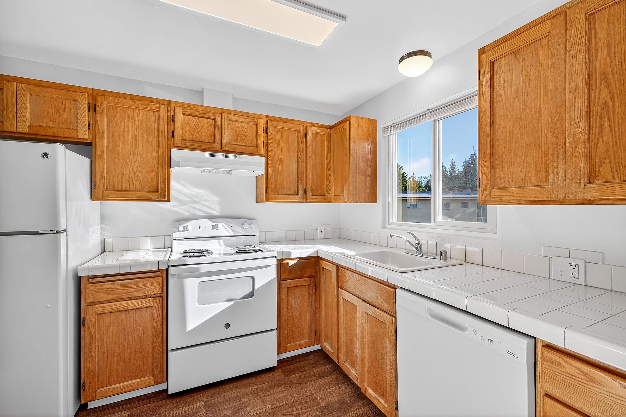 Bright and modern kitchen featuring wooden cabinets, a white stove and refrigerator, a sink under a window, and a dishwasher. The countertops are tiled, and there is ample natural light coming in, creating an inviting space for cooking and dining.