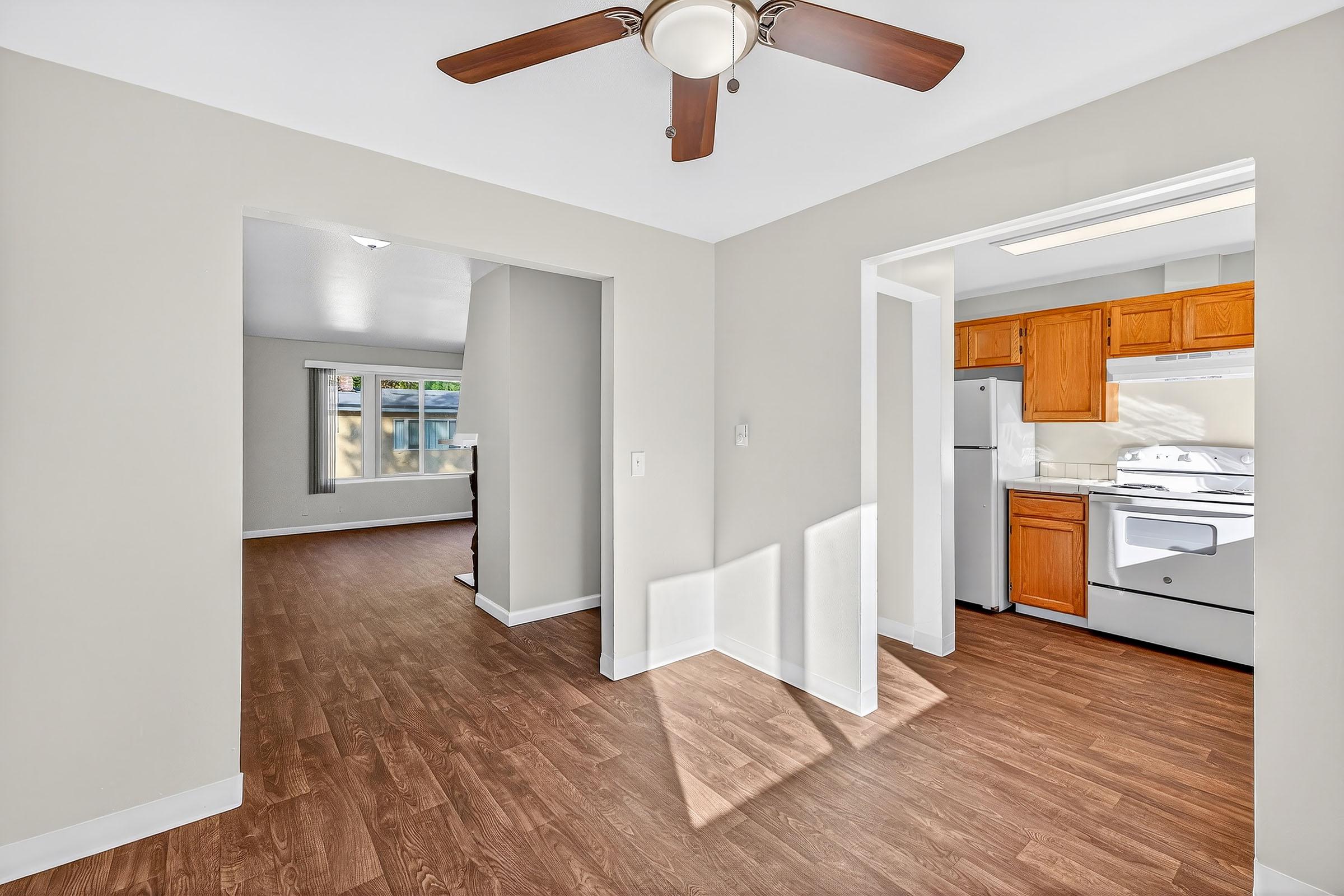 Bright interior of a home featuring an open layout. The living area has light-colored walls and a ceiling fan, while a kitchen with wooden cabinets is visible to the right. Natural light filters in, creating a warm and inviting atmosphere.