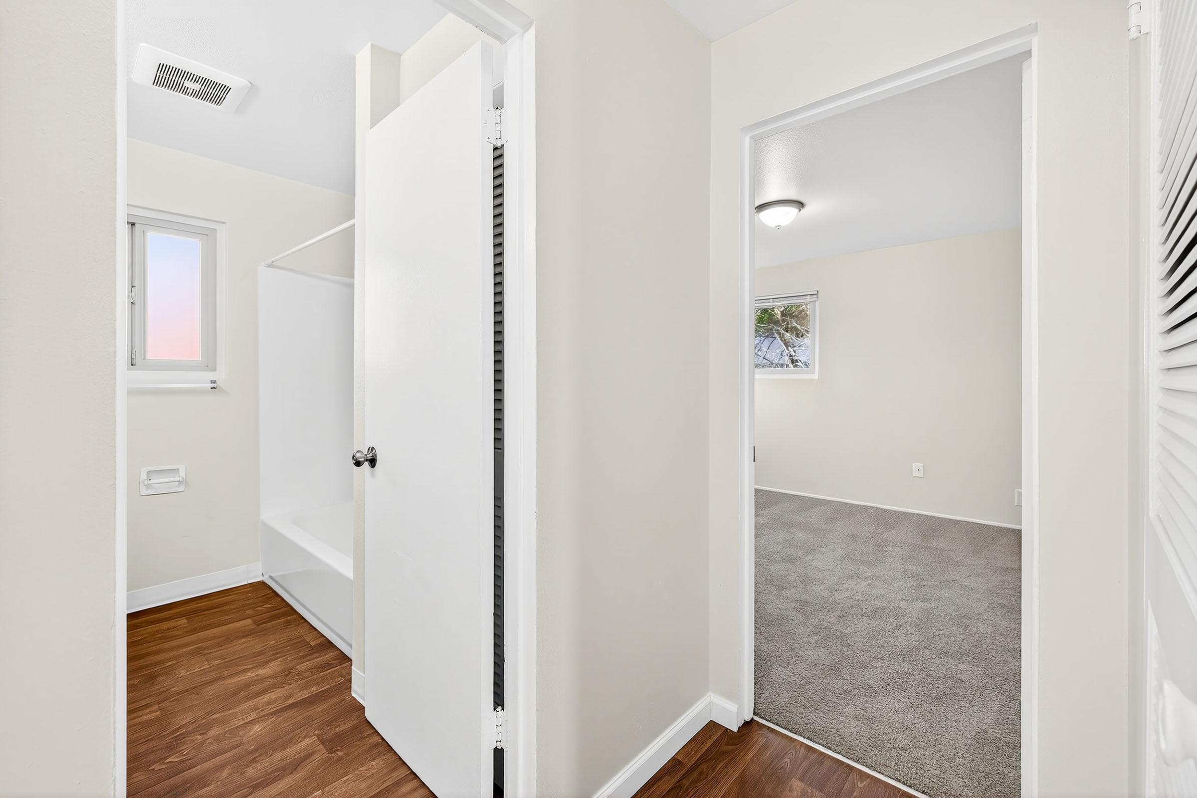 Interior view of a hallway in a home, featuring a doorway leading to a room with carpet and a window. To the left, there is a bathroom area with a shower and light-colored walls. The floors are hardwood, and there is a light fixture on the ceiling. Natural light is visible through the windows.