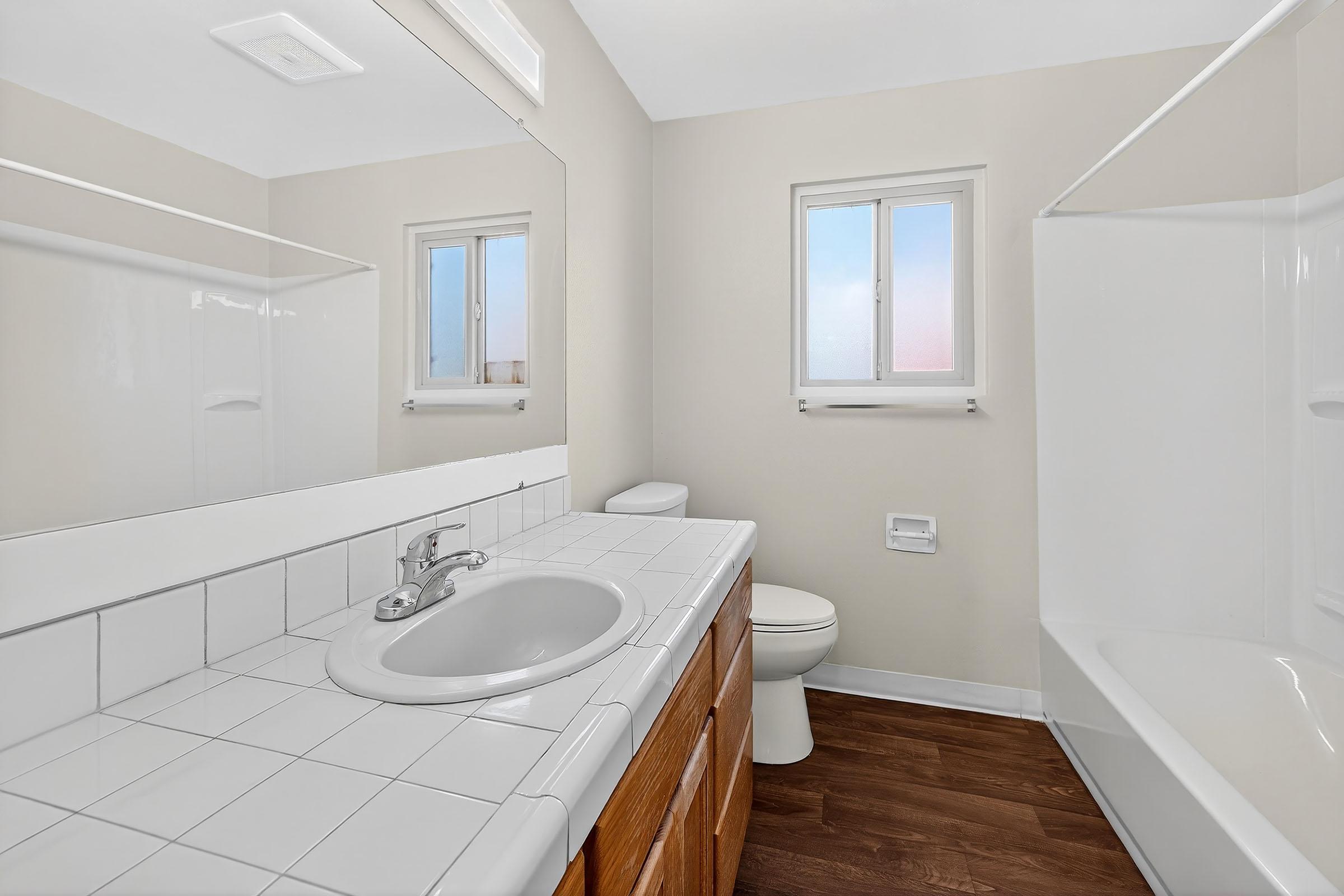 A clean and well-lit bathroom featuring a white tiled sink counter, a modern faucet, a white toilet, and a shower-tub combo. Two windows allow natural light to brighten the space, complemented by light beige walls and dark wooden flooring.