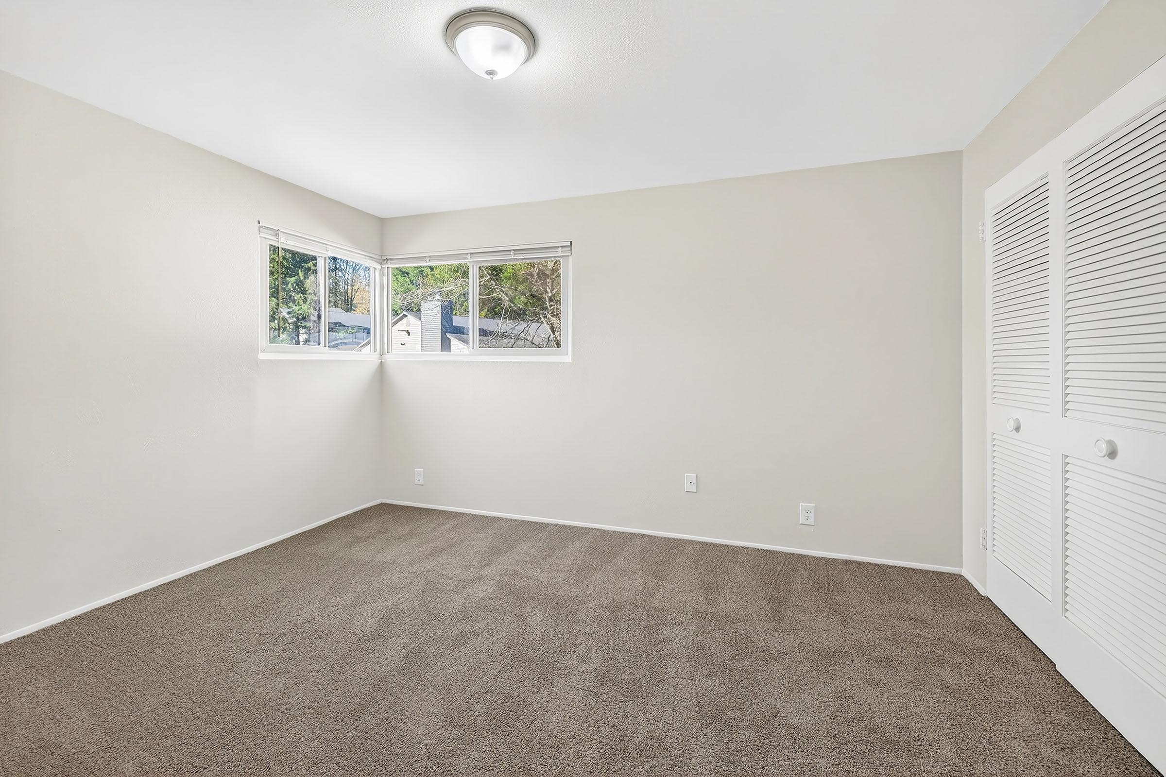 Empty room with light beige walls and carpeted flooring. Features a ceiling light fixture and two windows allowing natural light. One wall has double doors leading to a closet, and there are no furnishings or decorations present. The overall space feels bright and open.