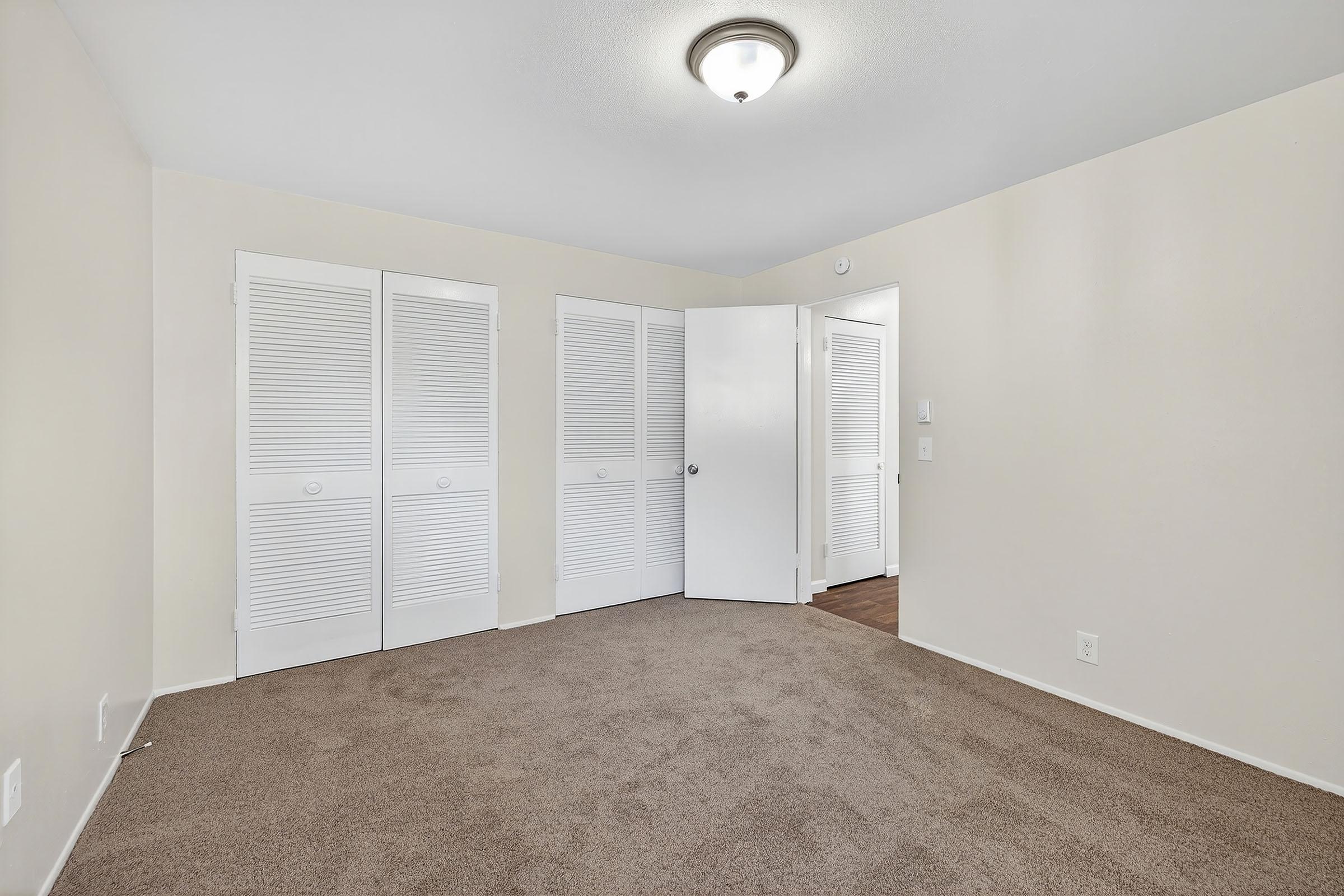 A well-lit, empty room with beige walls and carpet. There are three white, louvered closet doors on one wall, and a white door leading to another area. The ceiling features a simple light fixture, creating a clean and spacious atmosphere.